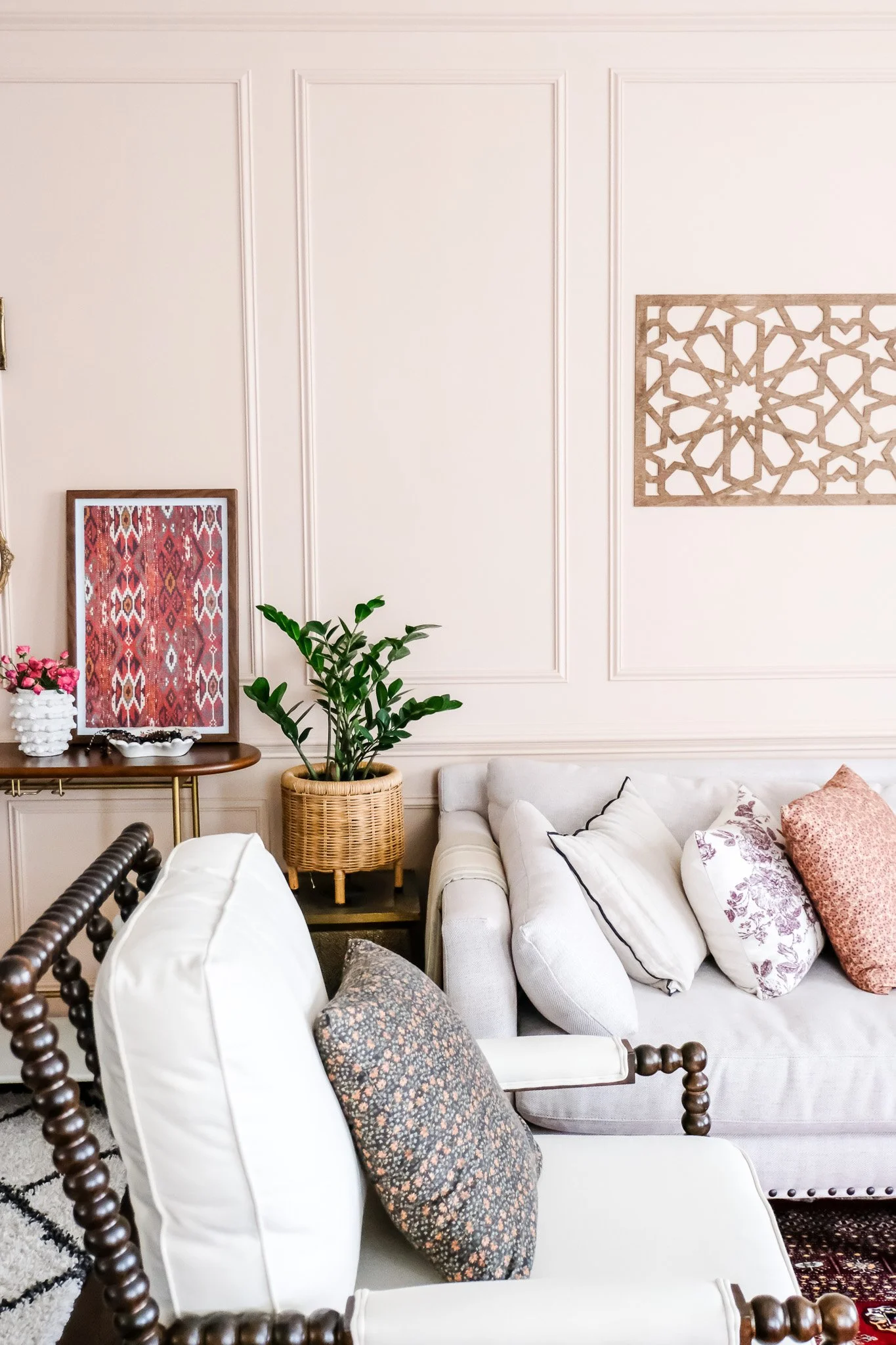 Interior living room with white walls, a beige sofa with patterned pillows, a dark wooden armchair with a floral pillow, a side table with a plant in a wicker basket, artwork on the walls, and decorative items.