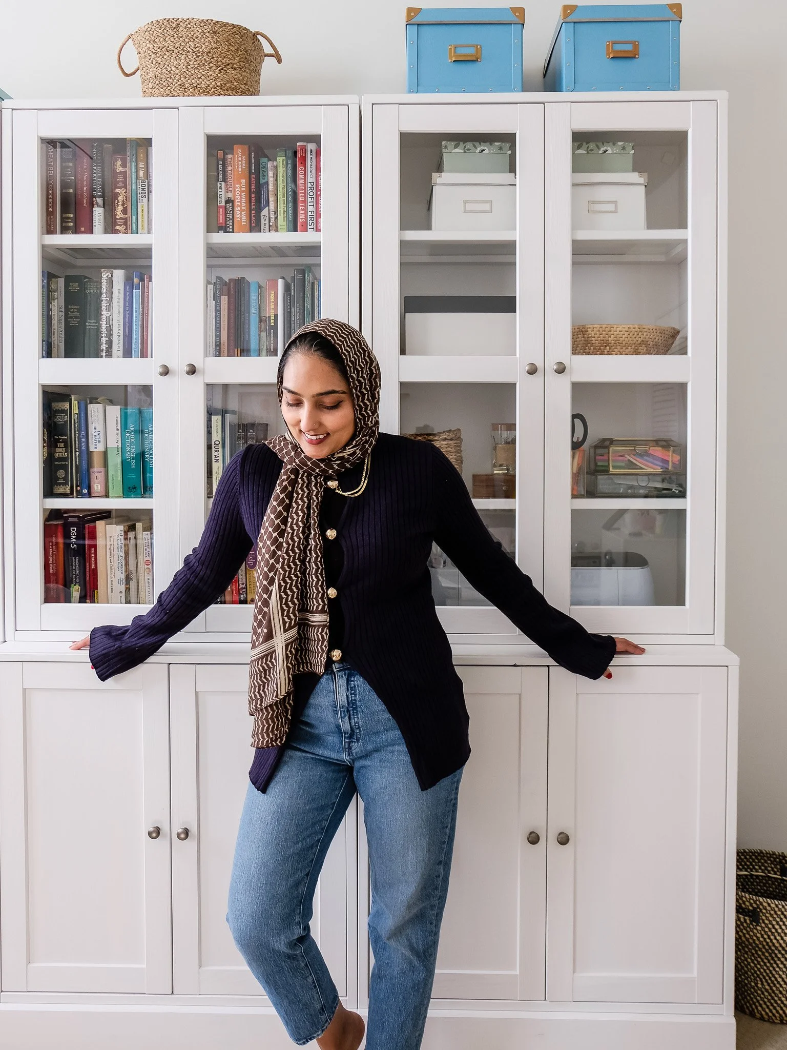 A woman wearing a patterned headscarf, dark sweater, and jeans standing in front of white cabinet with glass doors filled with books and storage boxes.