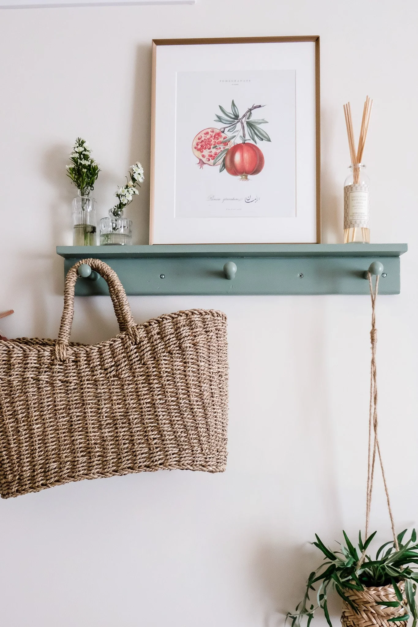 Decorative wall shelf holding a botanical print, small vases with flowers, a reed diffuser, a hanging potted plant, and a woven bag.