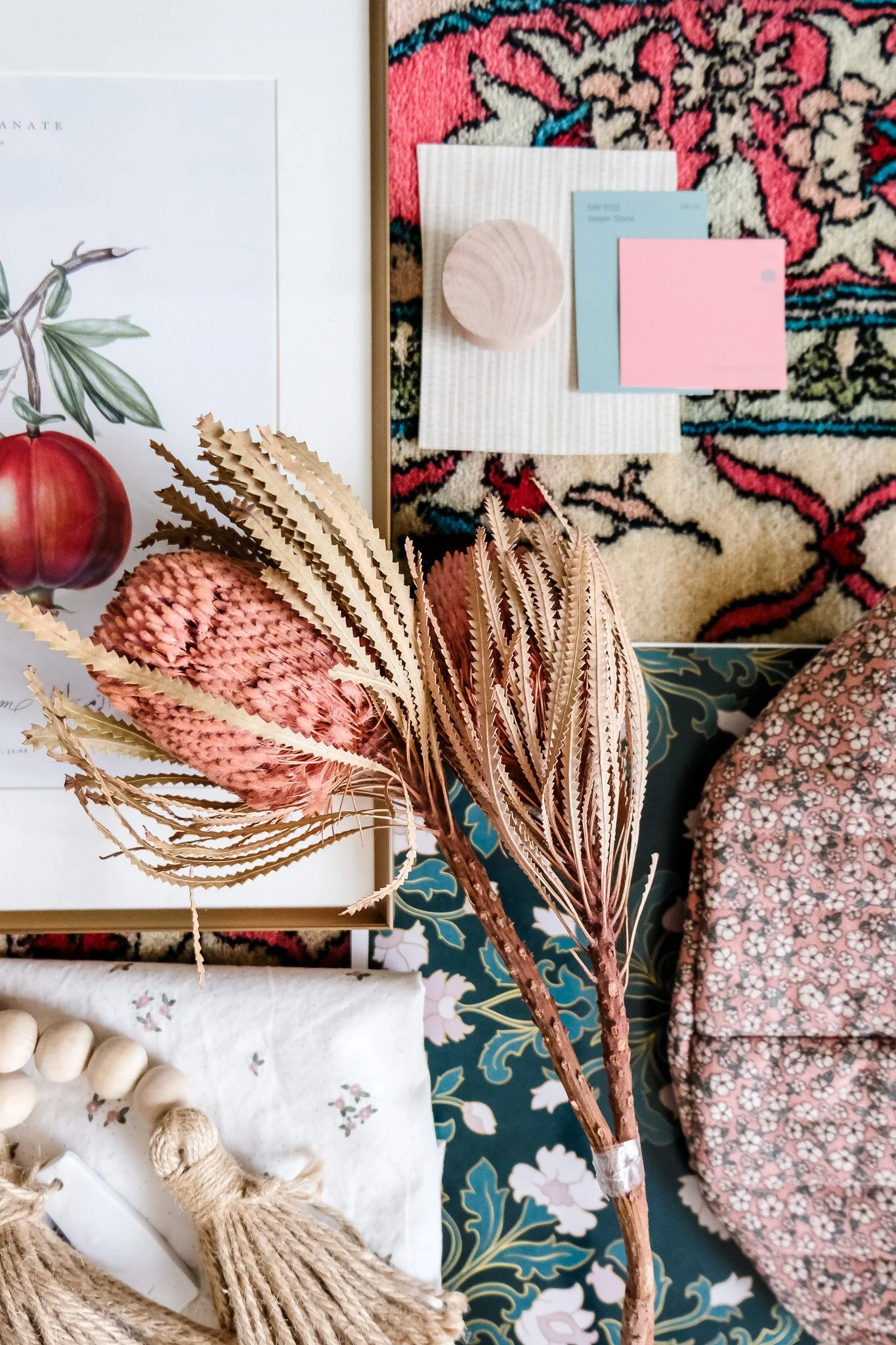 A tabletop with dried pink and beige faux foliage, a patterned floral bag, a beaded necklace with a tassel, and assorted stationery items including notepads, a wooden dish, and paper on a pink and blue patterned rug.