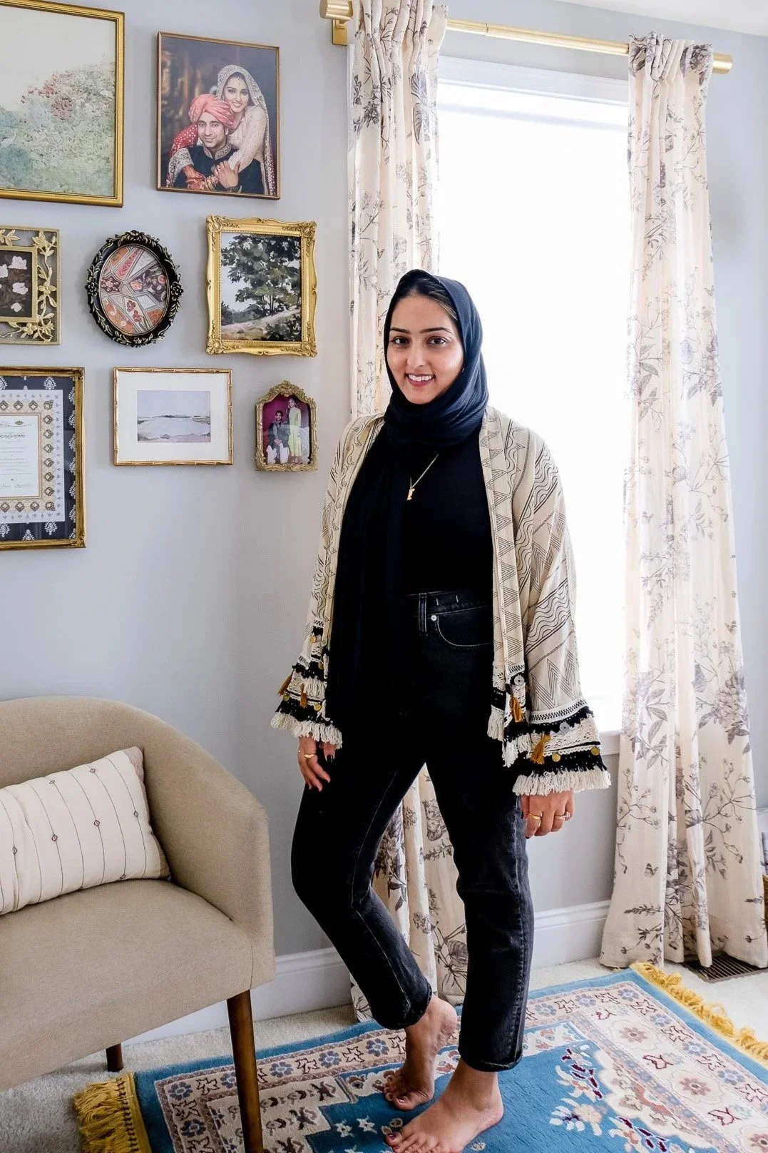 A woman with dark hair wearing a black scarf, a patterned beige jacket, and black jeans standing barefoot in a living room with light-colored walls and decorated with framed pictures and artwork, near a window with floral curtains and a beige couch.