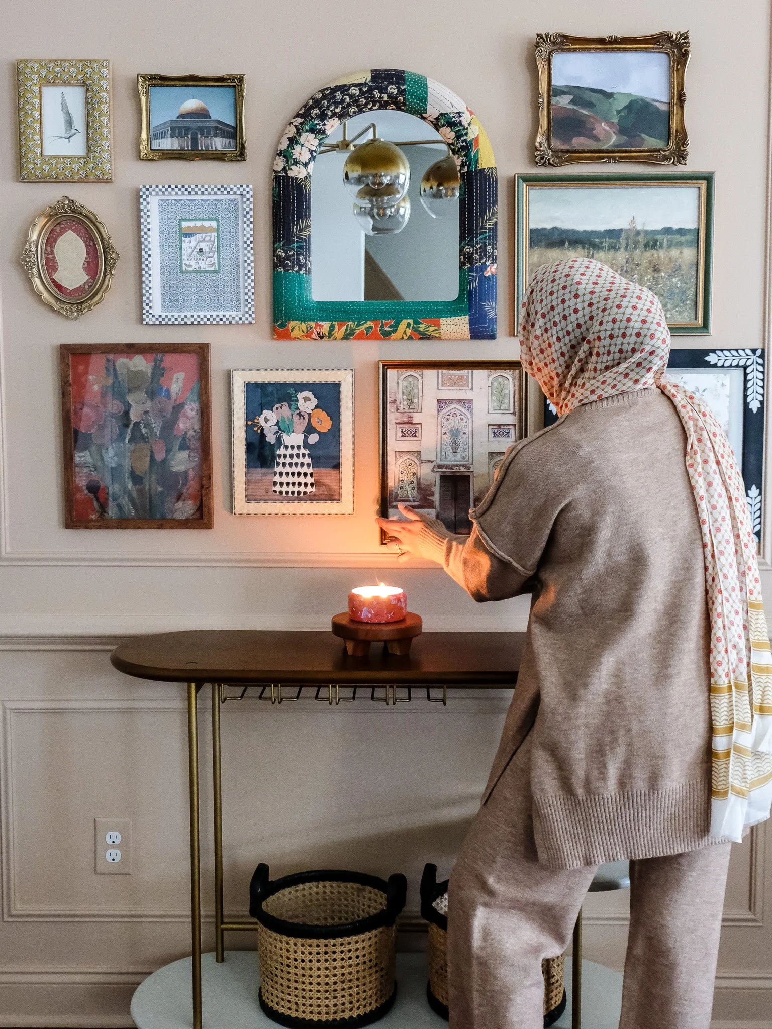 A person wearing a headscarf and casual clothing adjusts a framed picture on a wall decorated with various art prints and a mirror. There is a lit candle on a wooden table below, and two woven baskets underneath.