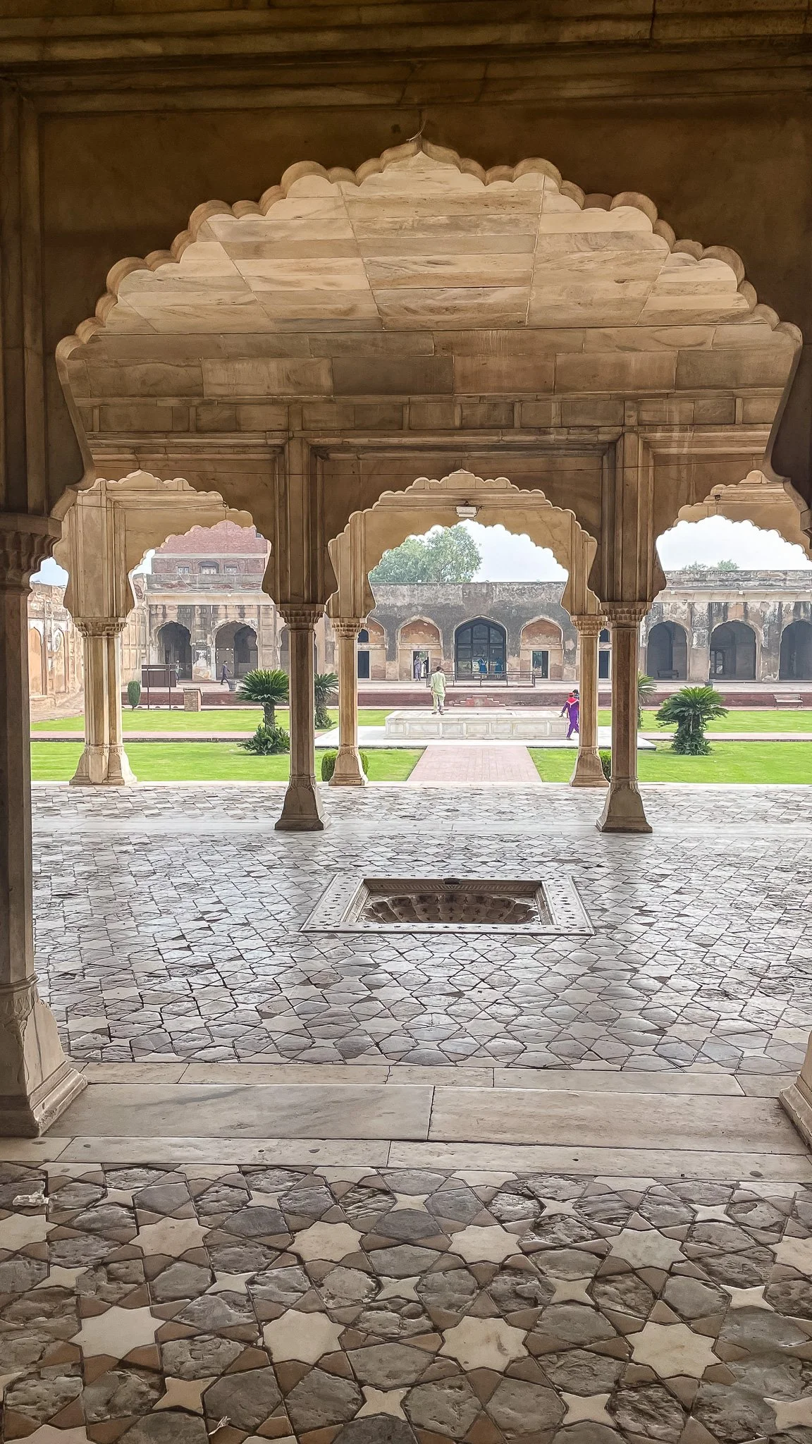 View of a historic Indian palace or fort with stone architecture, arched openings, marble floors, and surrounding gardens with small palm trees and people walking in the background.