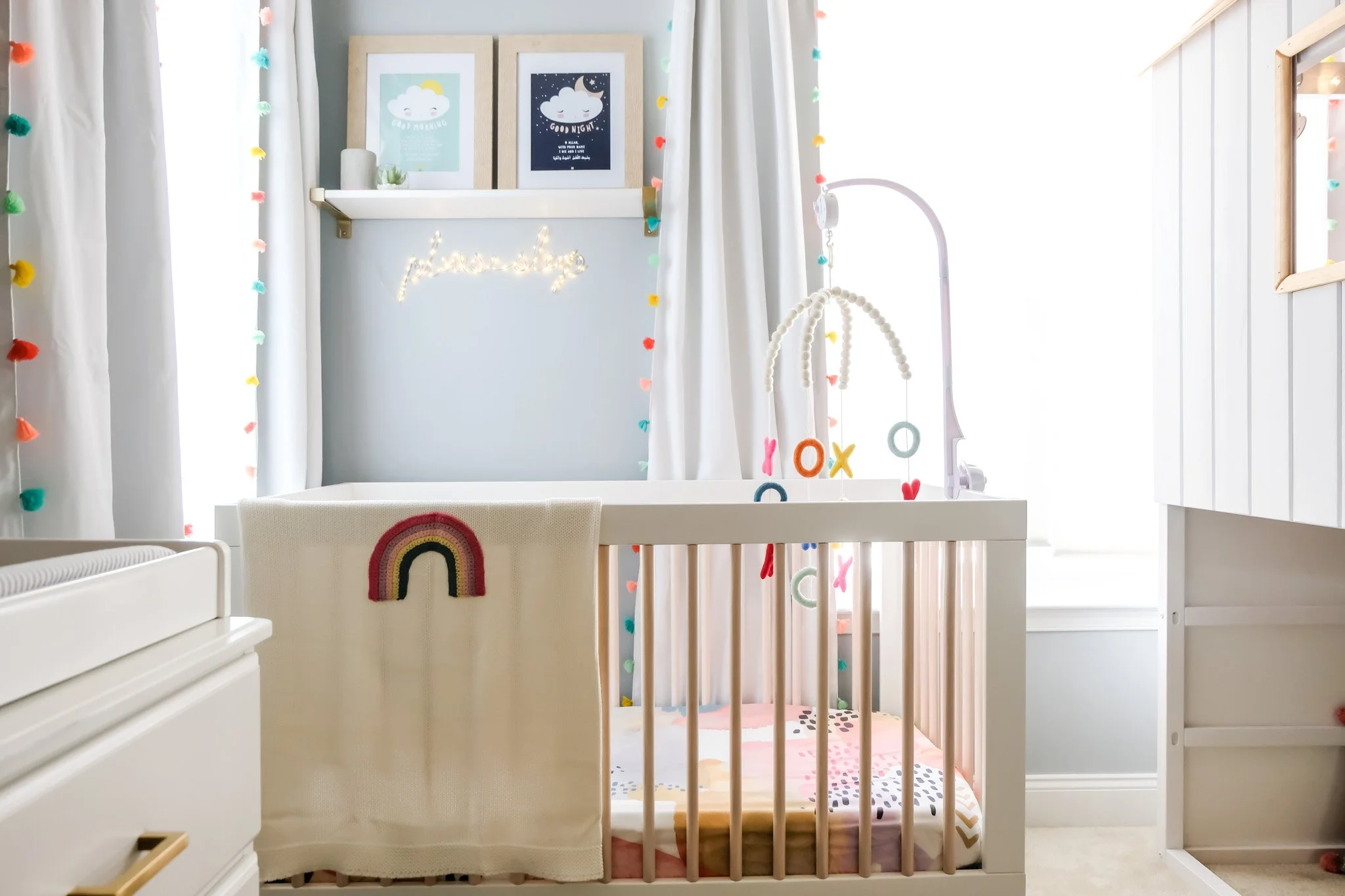 A nursery room with white cribs, a changing table, and a mobile hanging above the crib. The room has white curtains with colorful pom-pom trim, framed pictures on a shelf, and a neon sign that says 'flower' on a light blue wall.