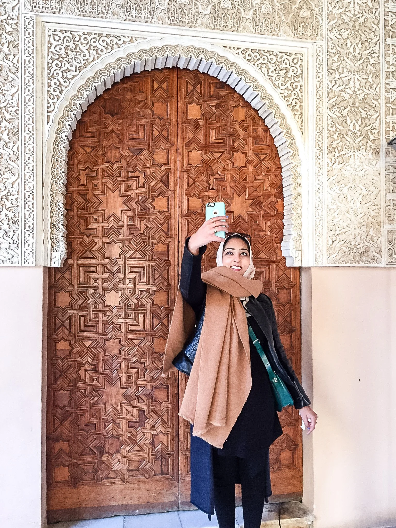 A woman taking a selfie in front of a wooden door with intricate Islamic geometric carvings. She is smiling and wearing a black outfit with a brown scarf and a headscarf, holding her phone up.
