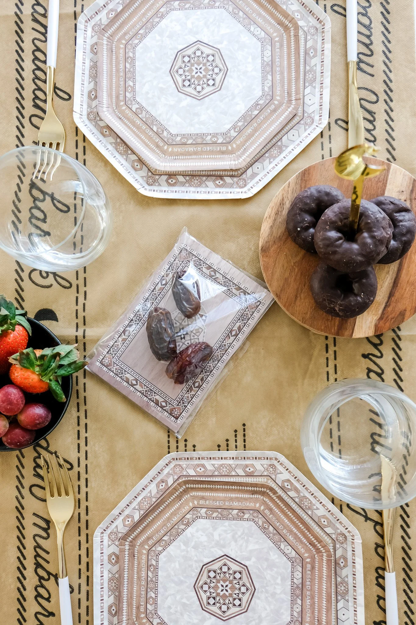 A table set for a meal with decorative tableware, including a patterned plate with the words 'Blessed Ramadan,' a glass of water, a bowl of strawberries and grapes, and a wooden serving board with chocolate-covered donuts and dates, with gold-colored utensils.
