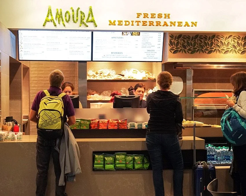 People ordering at a Mediterranean food counter named Amoura, with a menu board above, a display of baked goods behind the counter, and snacks on the counter.