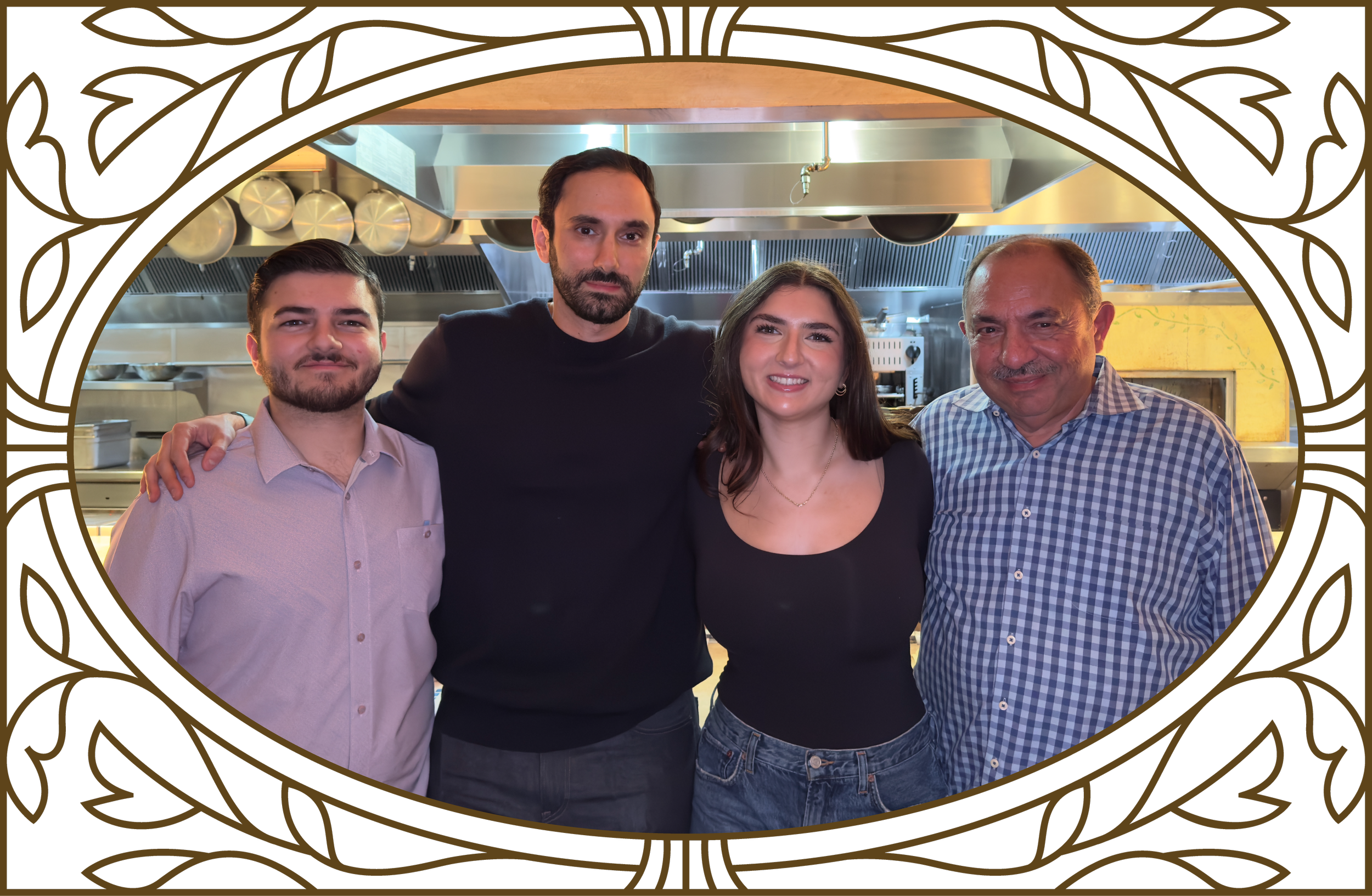 Four people standing together in a kitchen, smiling for the photo. The group consists of two men and two women, with the man in the middle having his arms around the others. The kitchen background features pots and stainless steel equipment.
