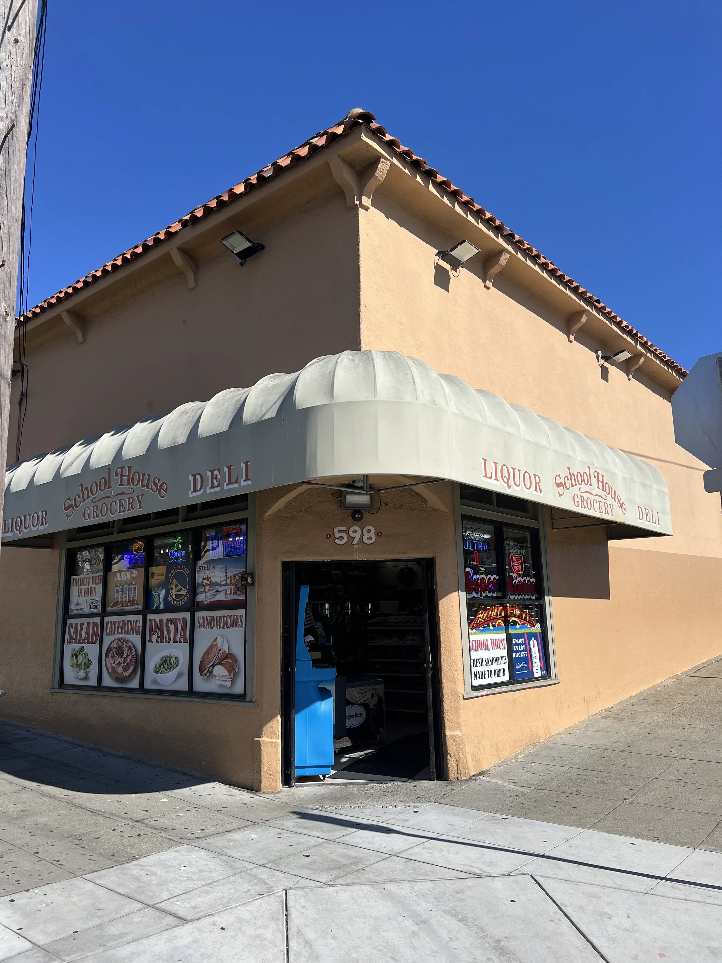 Facade of a corner store with a beige exterior wall, white awning with red lettering advertising liquor, school house, grocery, deli, and food items, and a large window displaying various posters and menus.