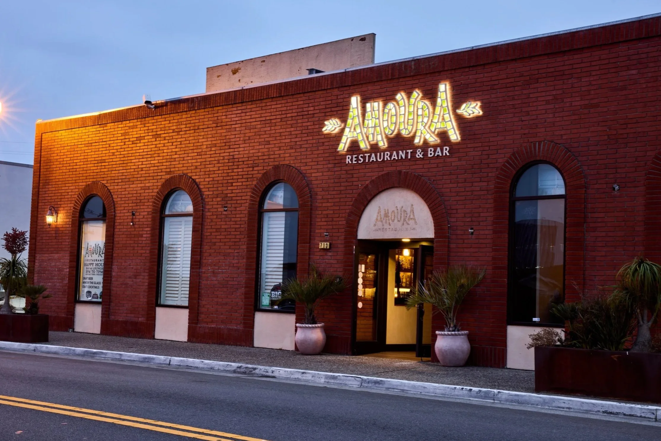 Brick building with illuminated sign that reads 'AMOUR RESTAURANT & BAR' and decorative leaves, large arched windows, two large potted plants outside, and street with double yellow lines in front.