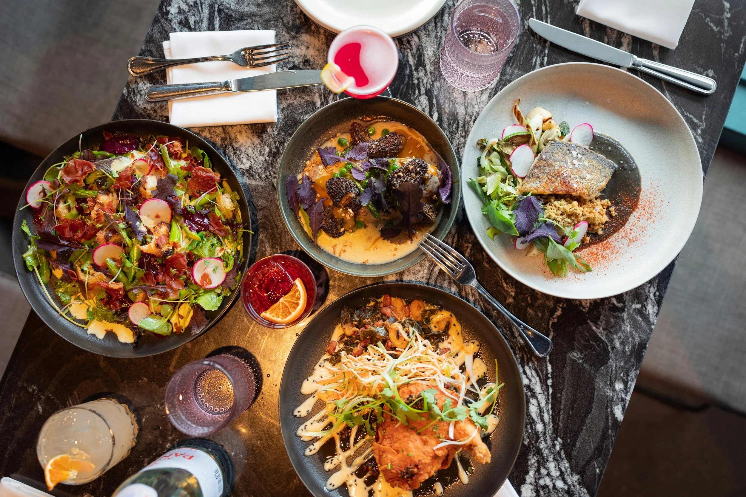 A table filled with various plates of food, including salads, a piece of cooked fish with salad, and other dishes, accompanied by drinks such as water and a pink cocktail with a lemon wedge, all on a dark marble surface.