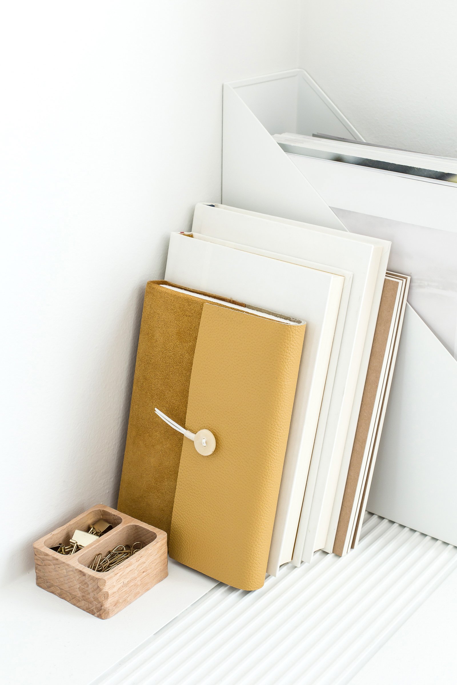 Collection of notebooks and folders arranged on a white surface near a window, with a small wooden container holding paper clips nearby.