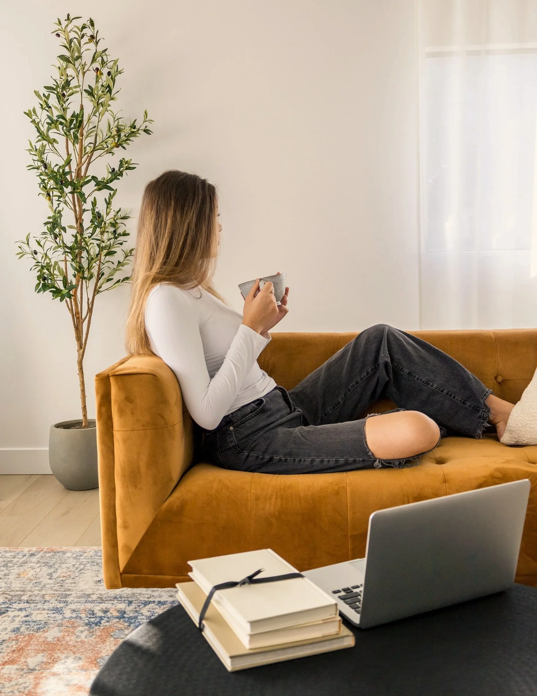 A woman with long light brown hair, wearing a white long sleeve shirt and ripped black jeans, sitting on an orange/brown sofa, holding a bowl and a spoon while looking at a window with sheer white curtains in a bright room with a potted plant, a coffee table with a laptop and notebooks.