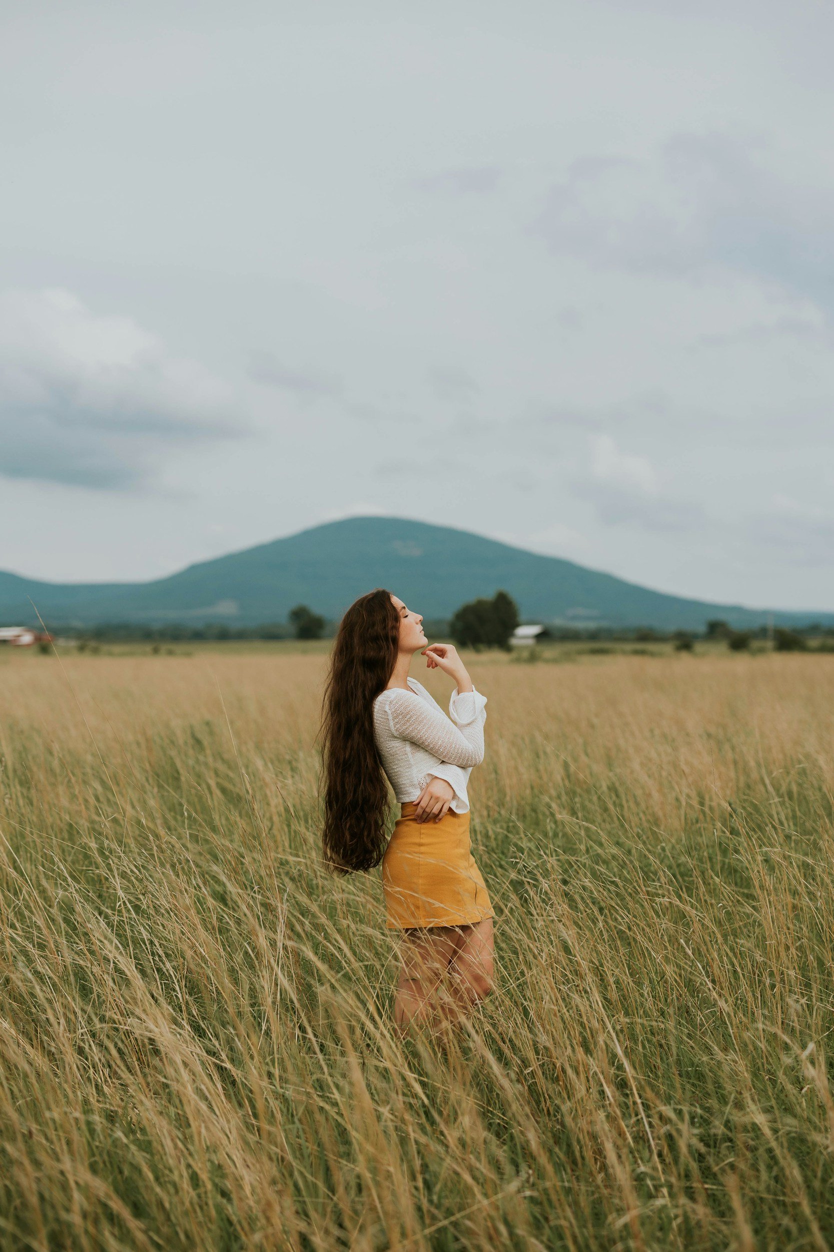 A woman with long brown hair standing in a field of tall grass during daytime, with her hand on her chin, looking contemplative, and mountains in the background.