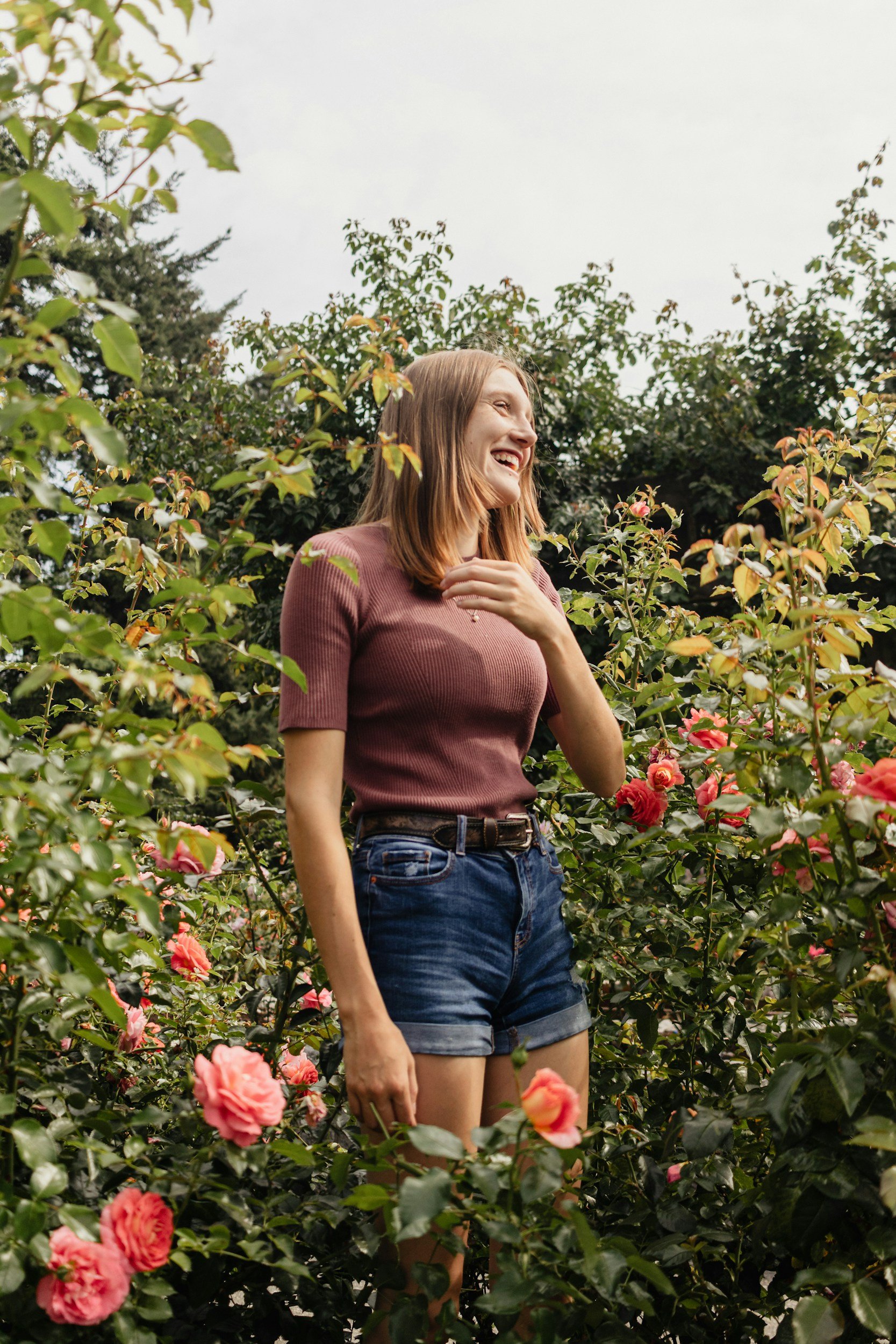 A young woman with shoulder-length red hair, wearing a maroon short-sleeve shirt and denim shorts, smiling and standing among pink roses in a garden.