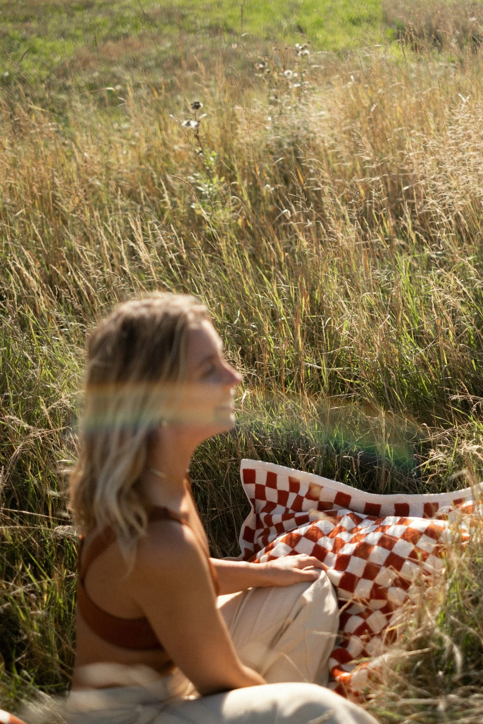 A woman sitting outdoors in a field of tall grass with a checkered blanket and a pillow, enjoying the sunlight on a sunny day.