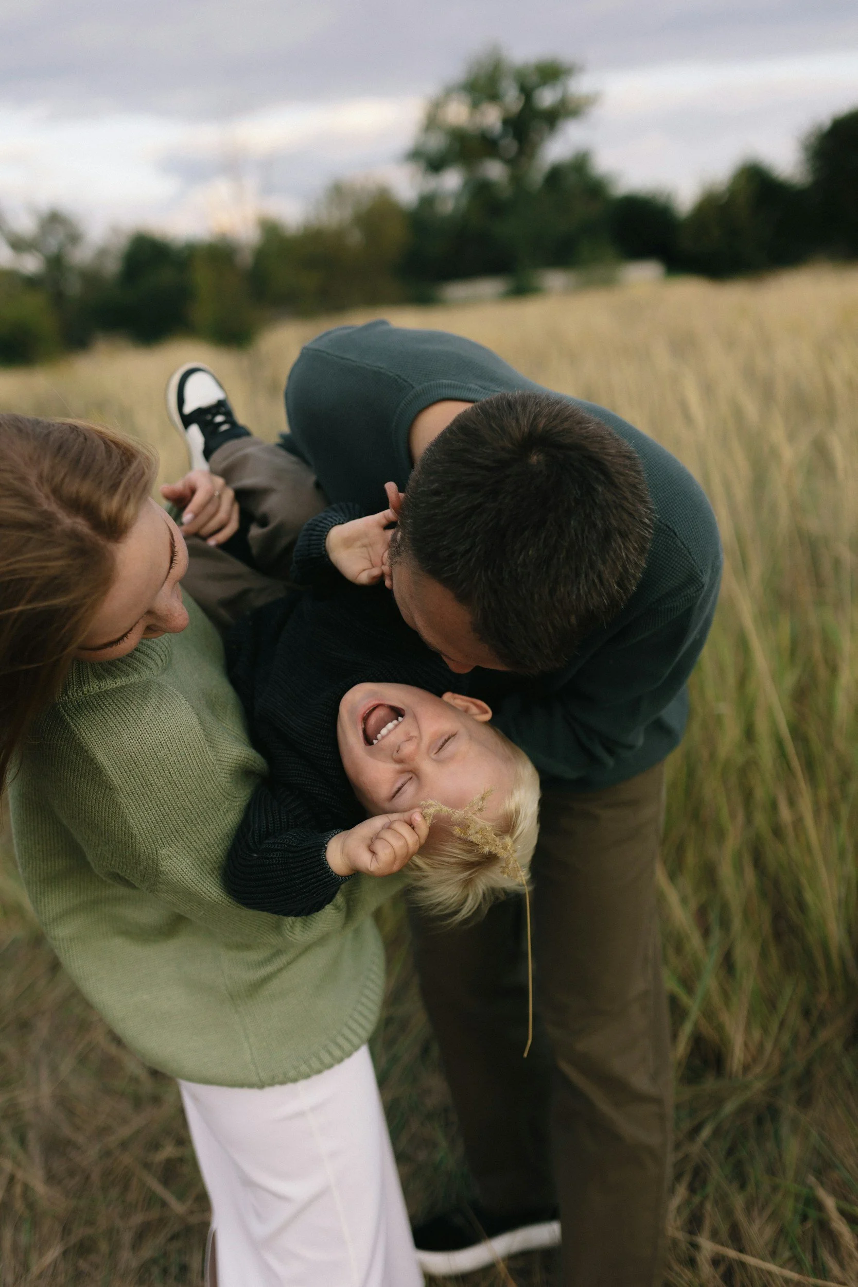 A family of three playing and laughing together outdoors in a field of tall grass during daytime.
