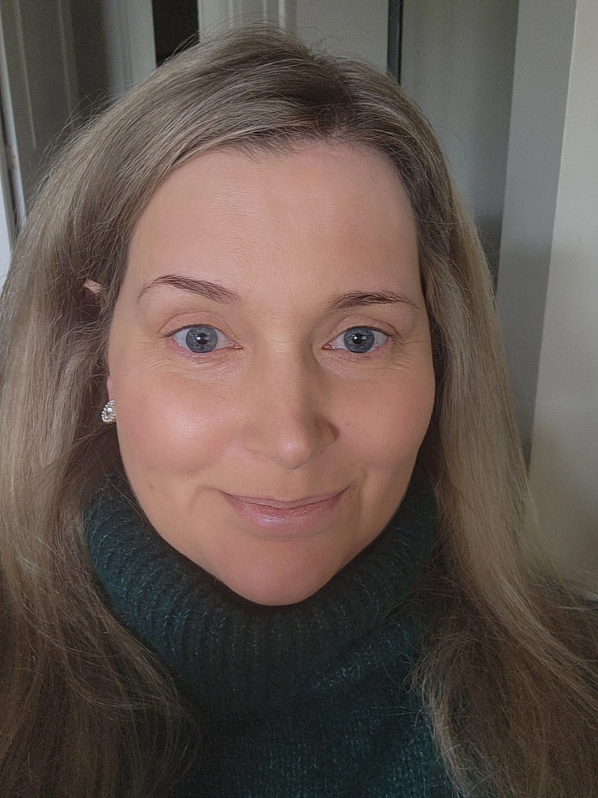 Close-up photo of a woman with light brown hair, blue eyes, wearing a black turtleneck sweater and a small earring, smiling softly, indoors.