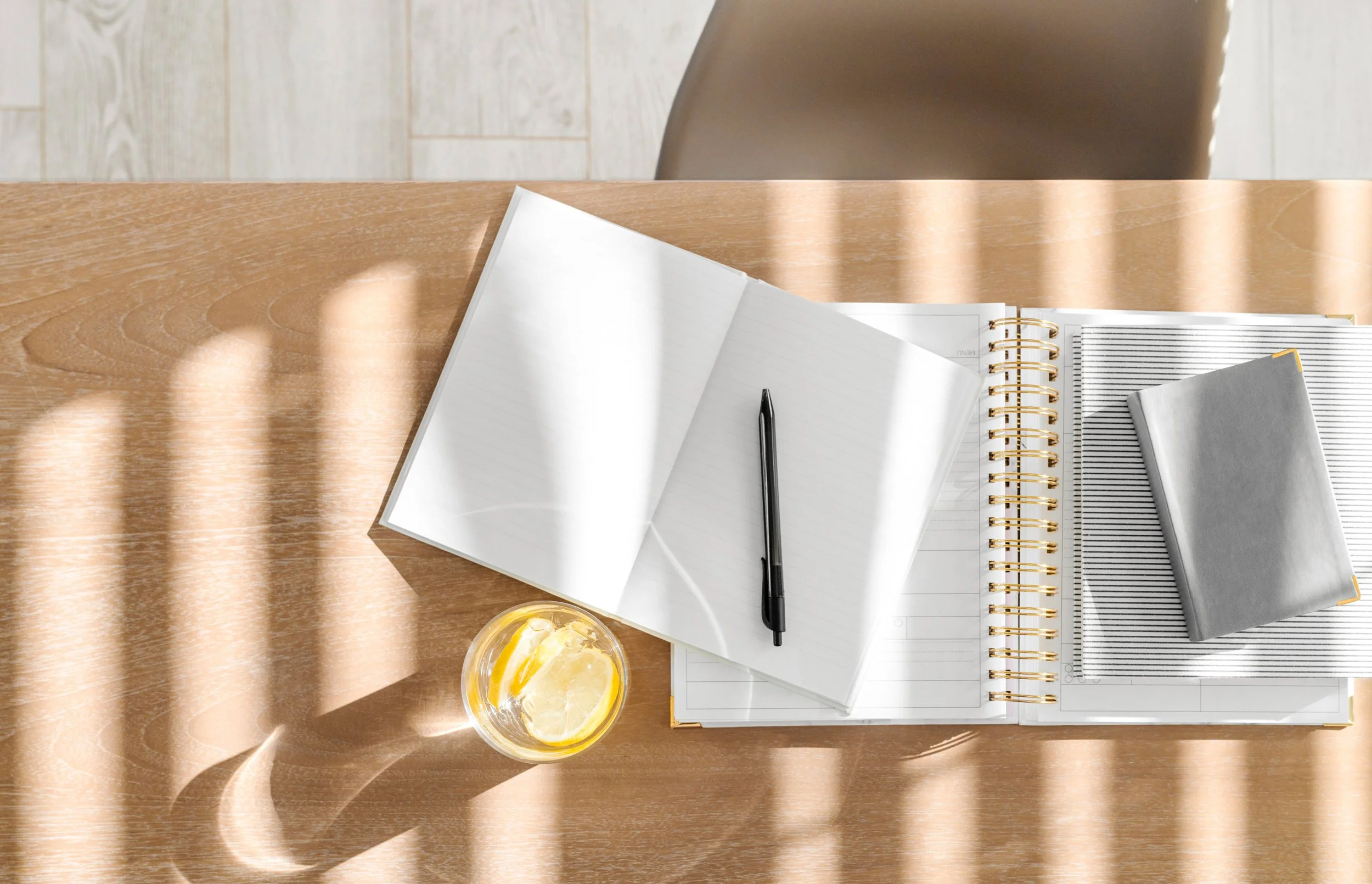 Overhead view of a wooden desk with an open notebook, a black pen, a closed notebook, a striped notepad, and a glass of lemon water, with sunlight casting striped shadows.