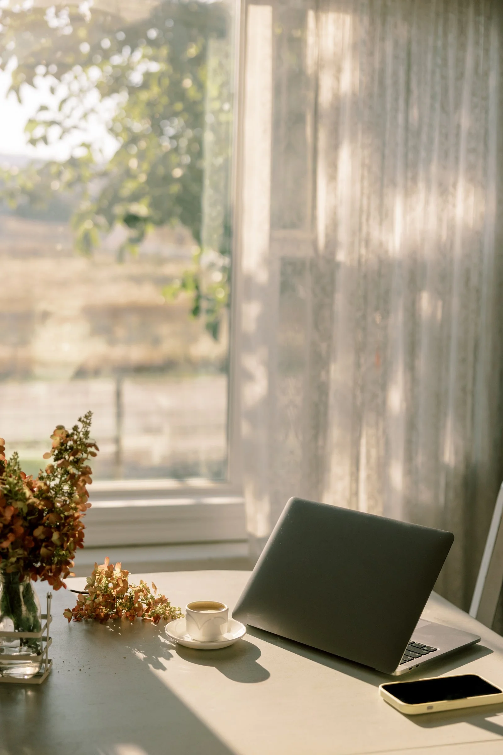 A desk near a window with sunlight streaming through, featuring a closed laptop, a smartphone, a cup of coffee on a saucer, and a vase of flowers.