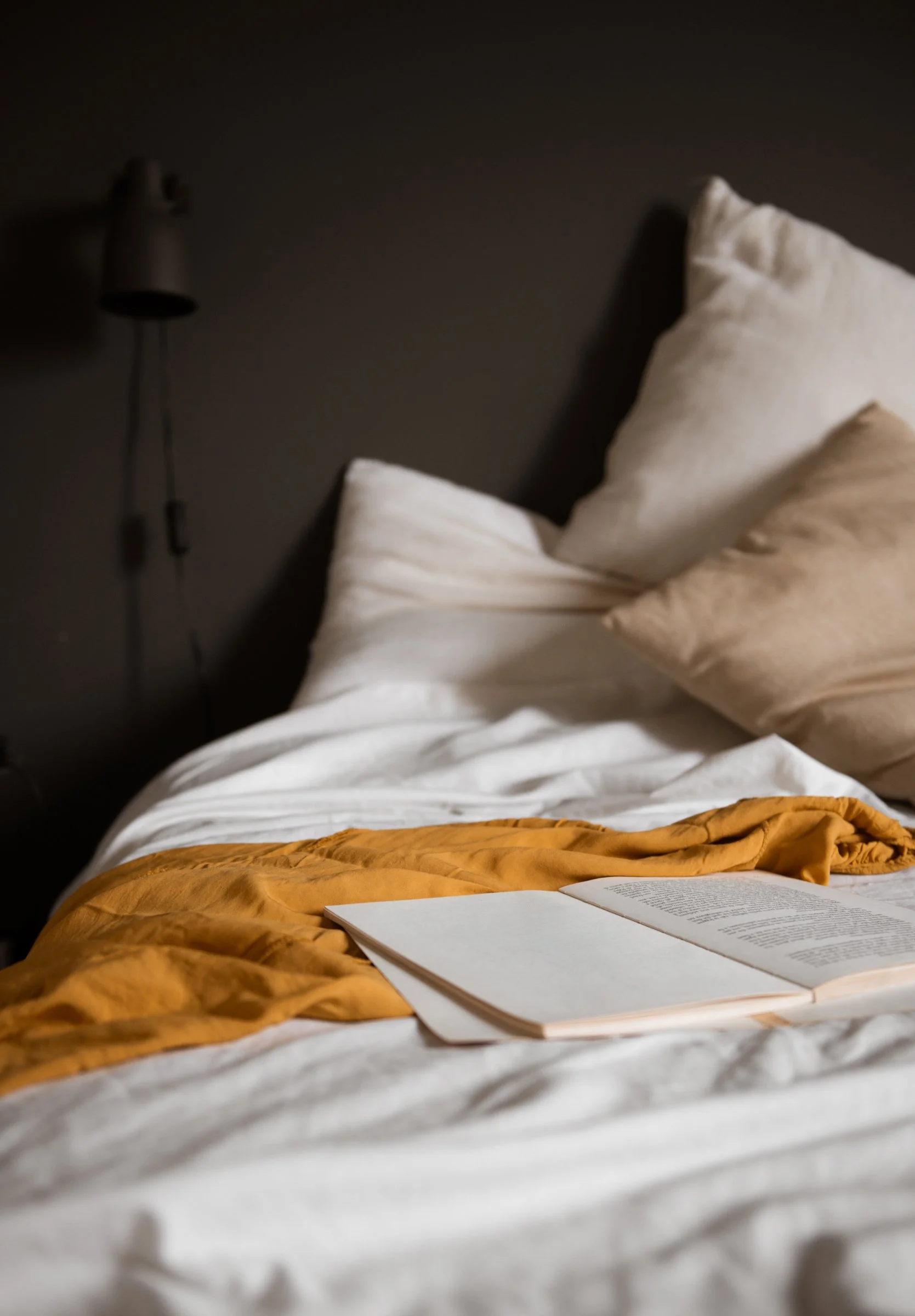 A bed with white sheets, beige pillows, and a mustard yellow blanket with an open book resting on it, against a dark wall with a wall-mounted lamp.