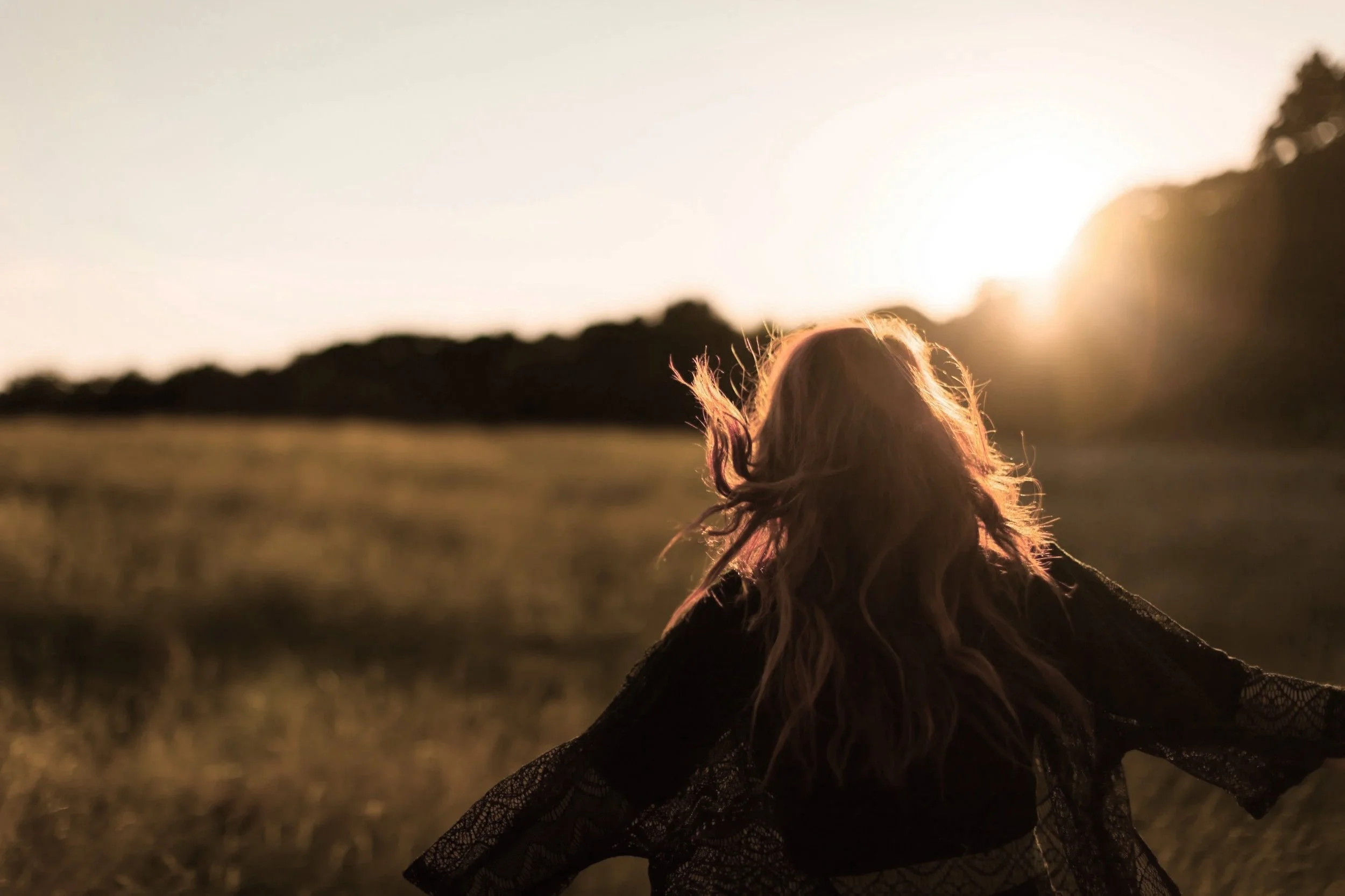 A woman with wavy hair standing in an open field during sunset, facing away from the camera, with her arms outstretched.