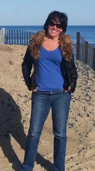 Author TL Cole, with sunglasses, and a smile, wearing a blue shirt, jeans, and a black jacket with fur trim, standing on sandy beach near the ocean.