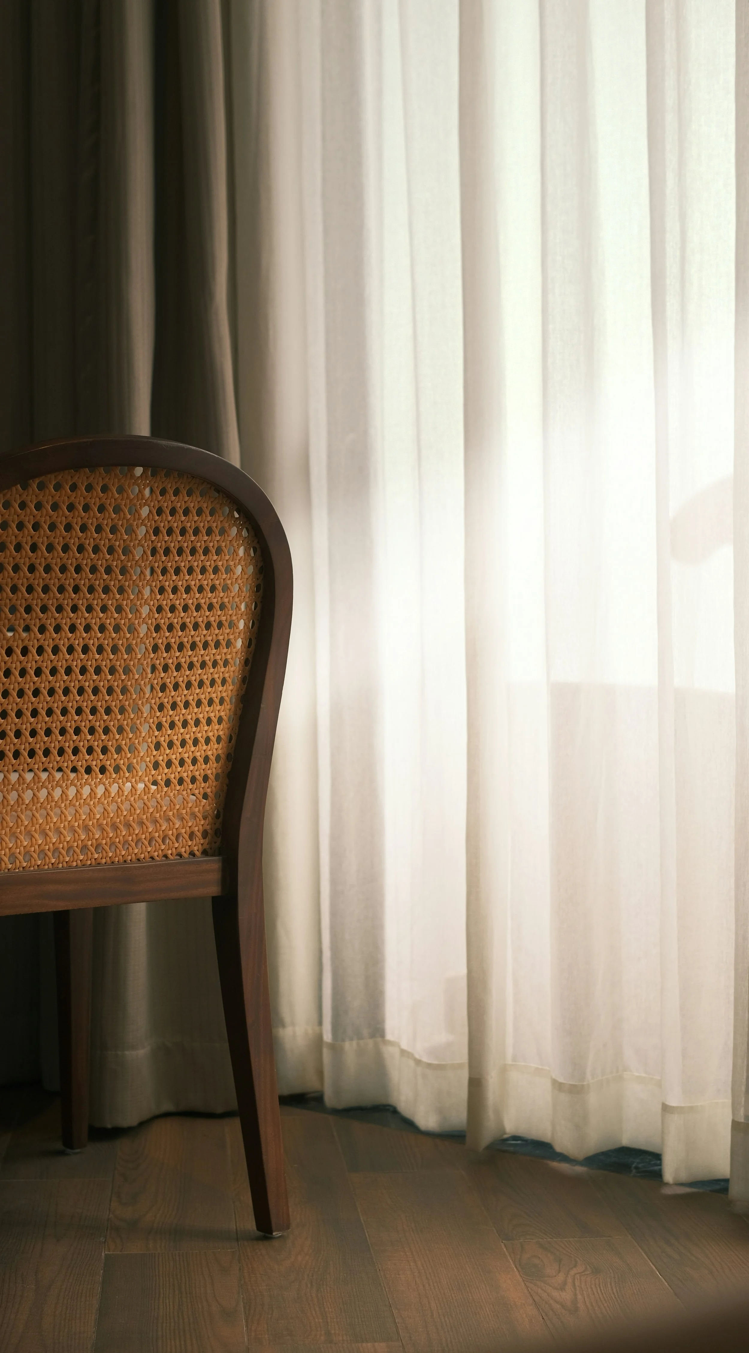 Part of a wooden chair with a rattan backrest, placed near sheer cream-colored curtains and a wooden floor in a minimalist interior..