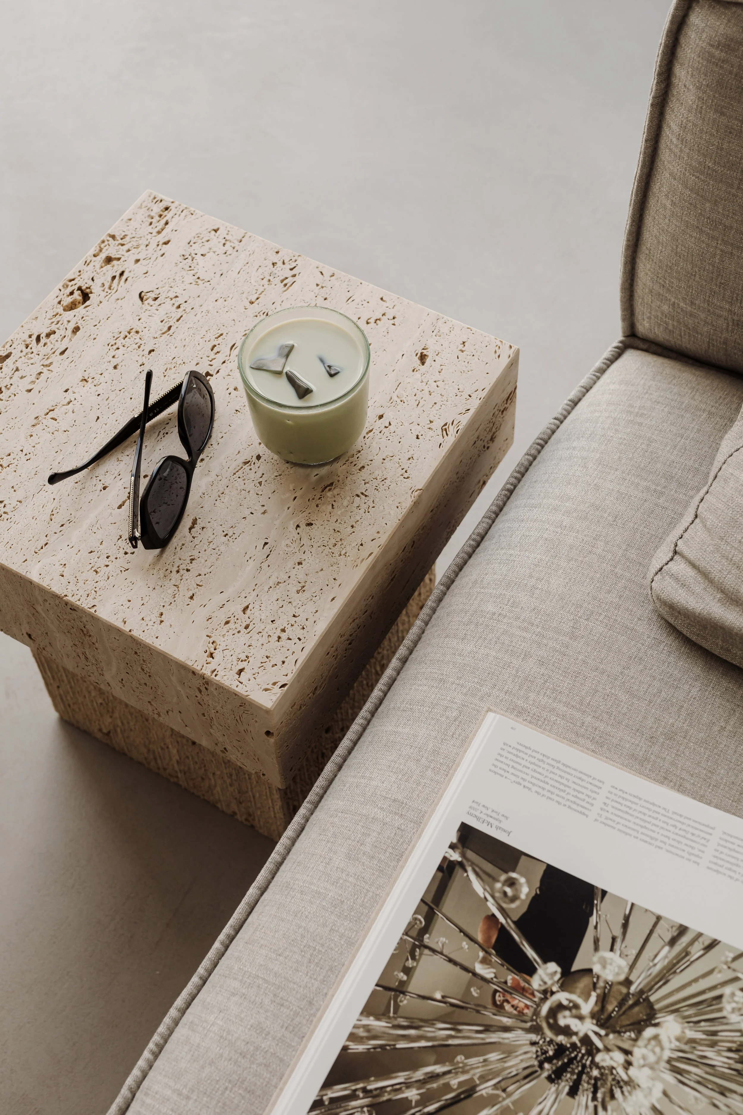 A  minimalist, beige, contemporary travertine side table with sunglasses and a matcha latte on top, positioned next to a light gray contemporary upholstered sofa with an open book resting on its cushion.