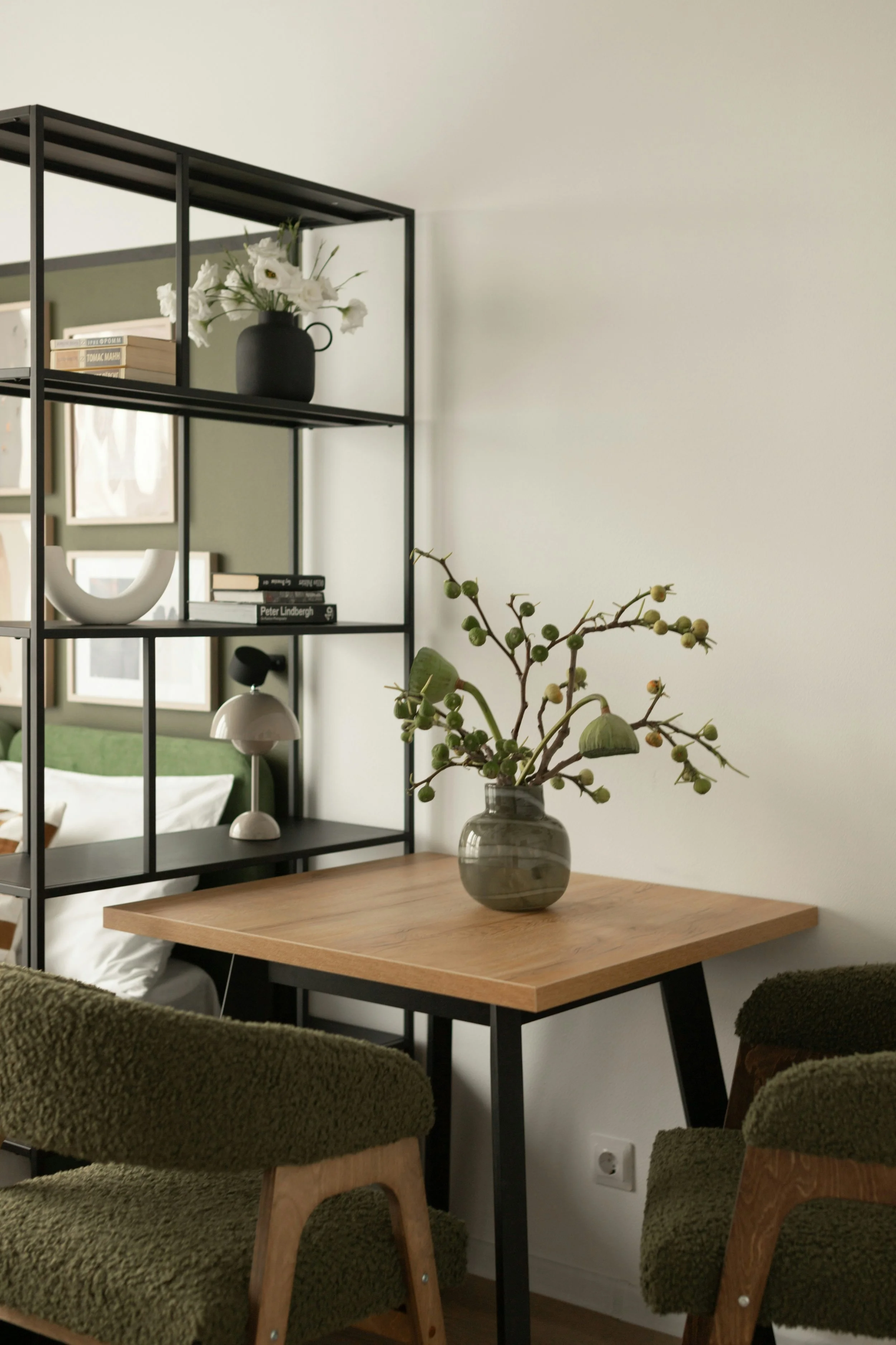 Interior of a modern living space featuring a black metal shelving unit with decorative items including a vase with white flowers, a pile of books, and a white sculpture. A wooden table with a greyish-green vase containing branches and green buds is in front of the shelving. There are plush green chairs with wooden arms surrounding the table, and a window with framed art hanging on the wall in the background.
