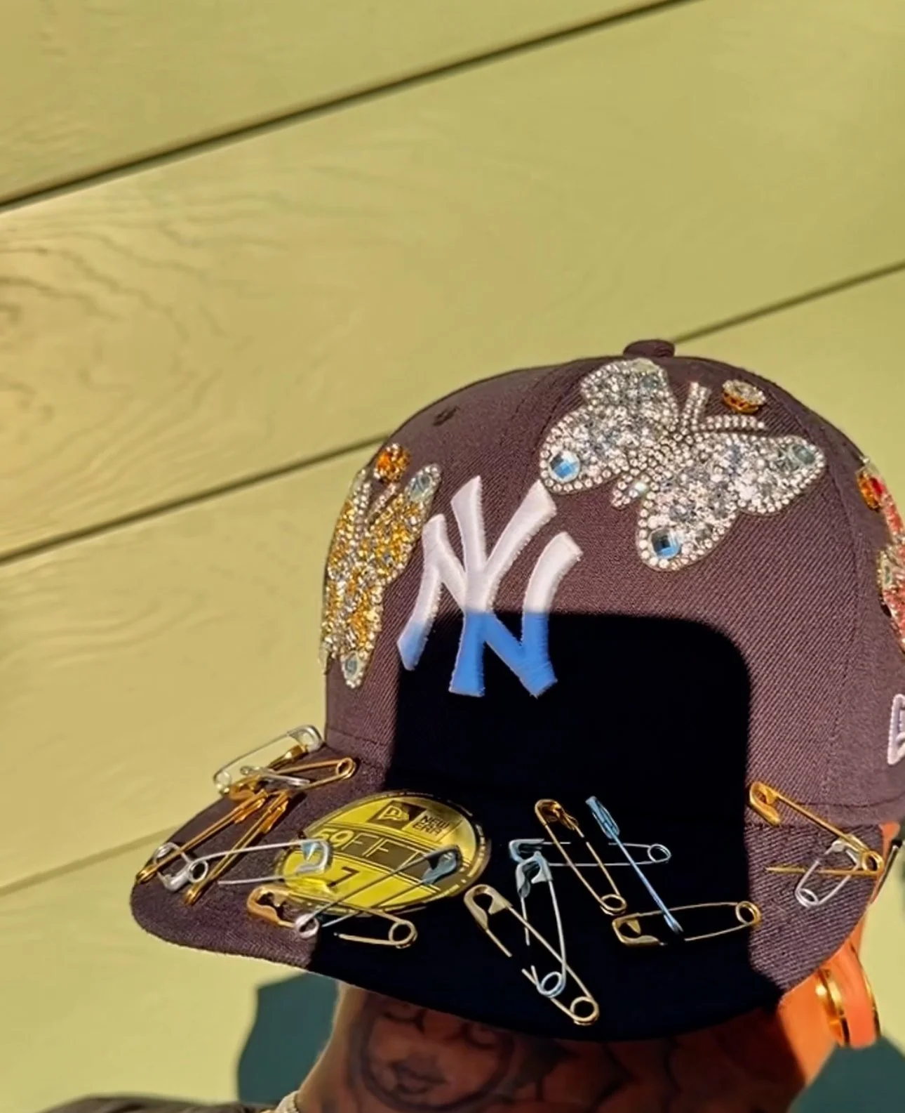 Classic yankee fitted cap decorated with rhinestone and embroidery patch designs, including a butterfly, bees, and the New York Yankees logo, with safety pins attached to the brim.