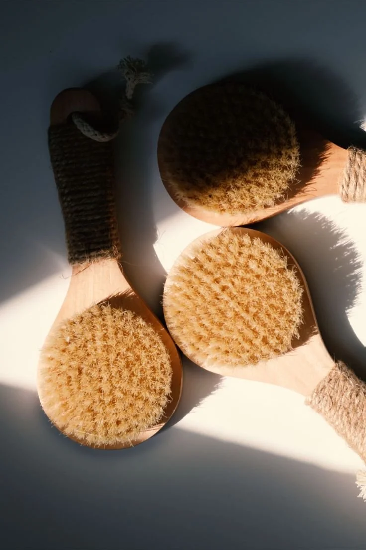Three wooden bath brushes with natural bristles and braided handles, arranged on a white surface with sunlight casting shadows.
