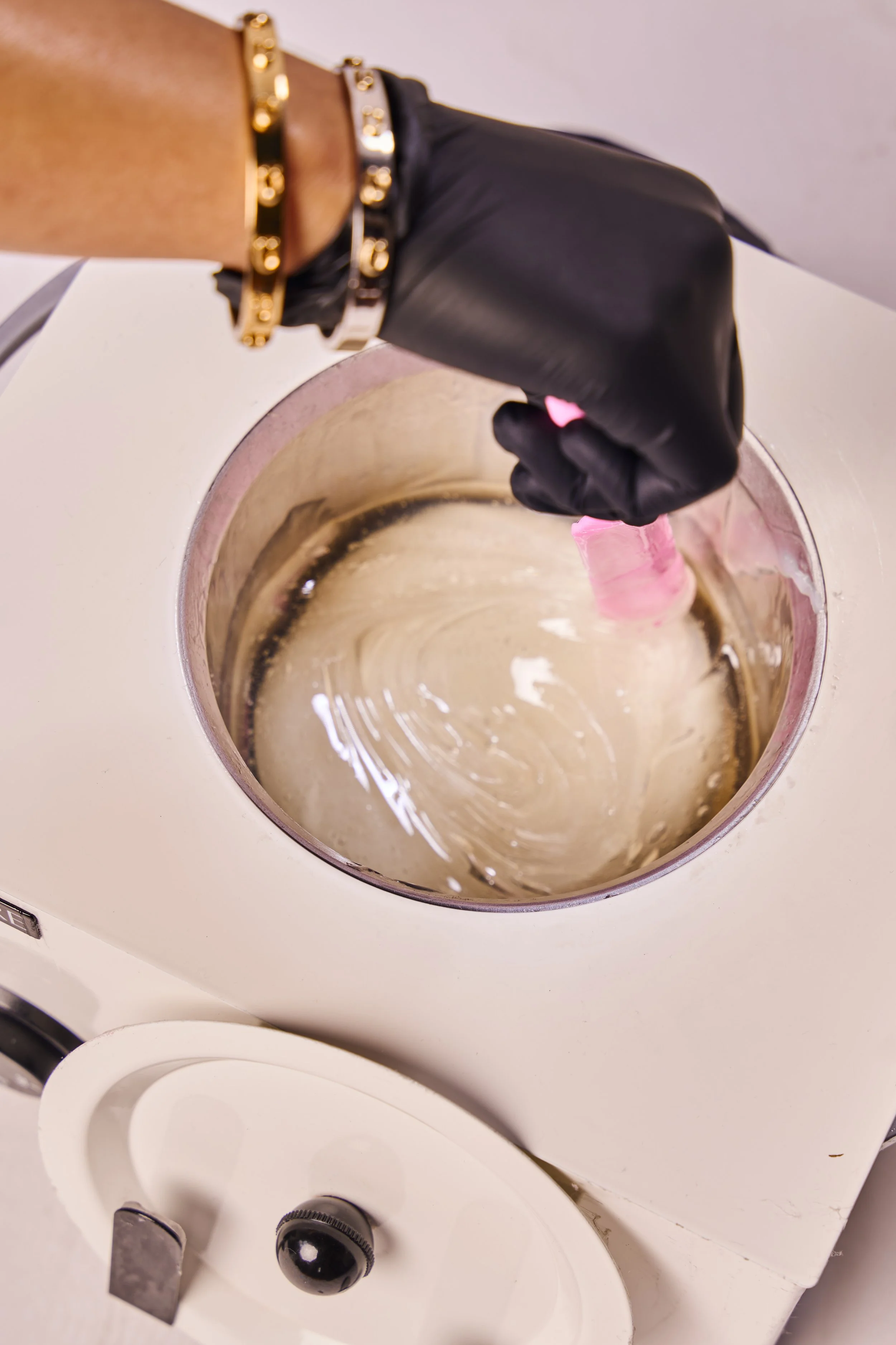 A person wearing black gloves and gold bracelets is holding a pink object over a large wax pot.