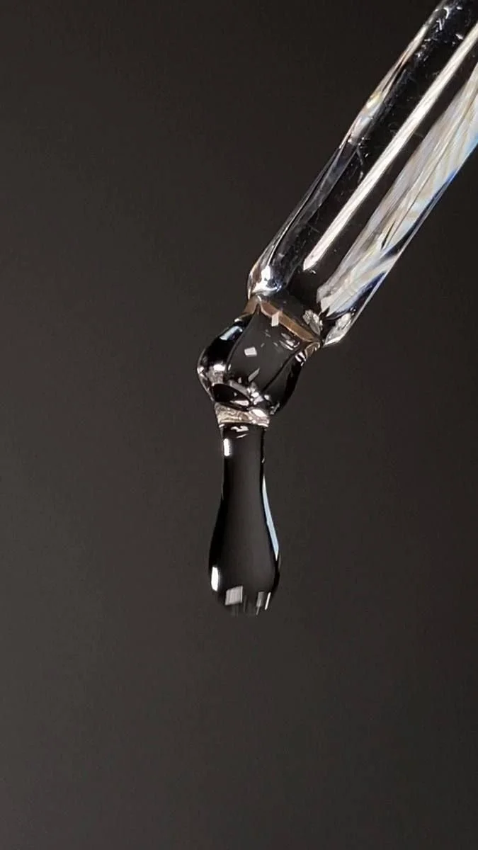 Close-up of a glass pipette releasing a droplet of black liquid against a dark background.