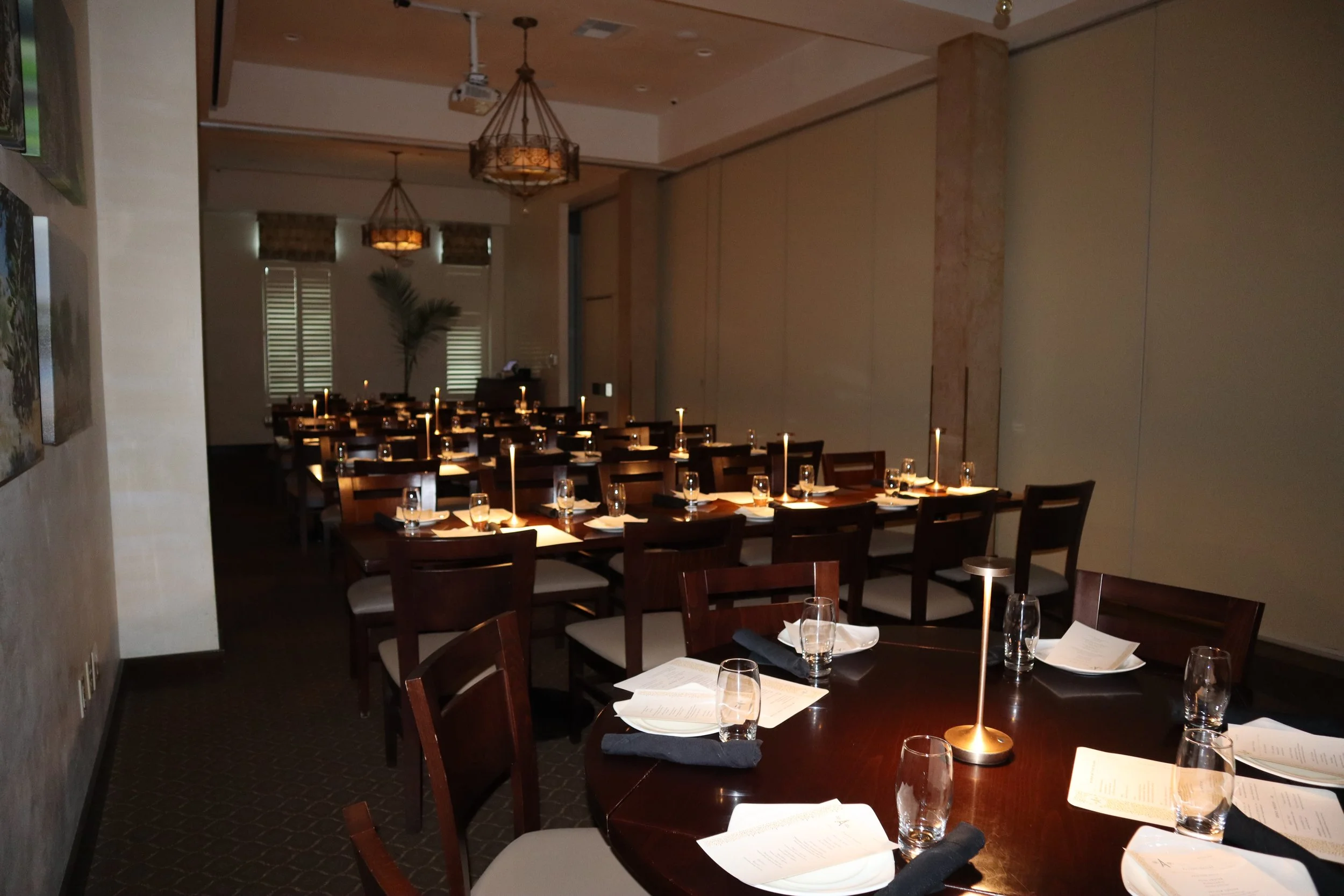 Elegant restaurant interior decorated for dinner with candlelit tables, wooden chairs, and soft lighting from chandeliers.
