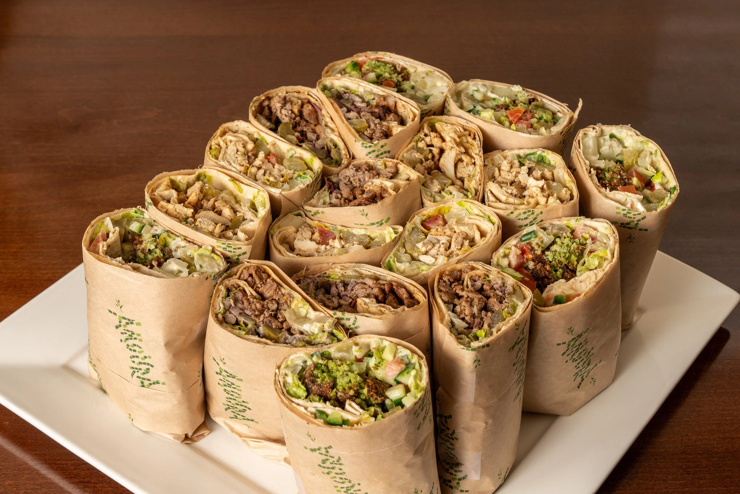 A plate of multiple wraps or burritos, sliced open to reveal beef, vegetables, and possibly cheese inside, wrapped in paper and arranged on a white platter.
