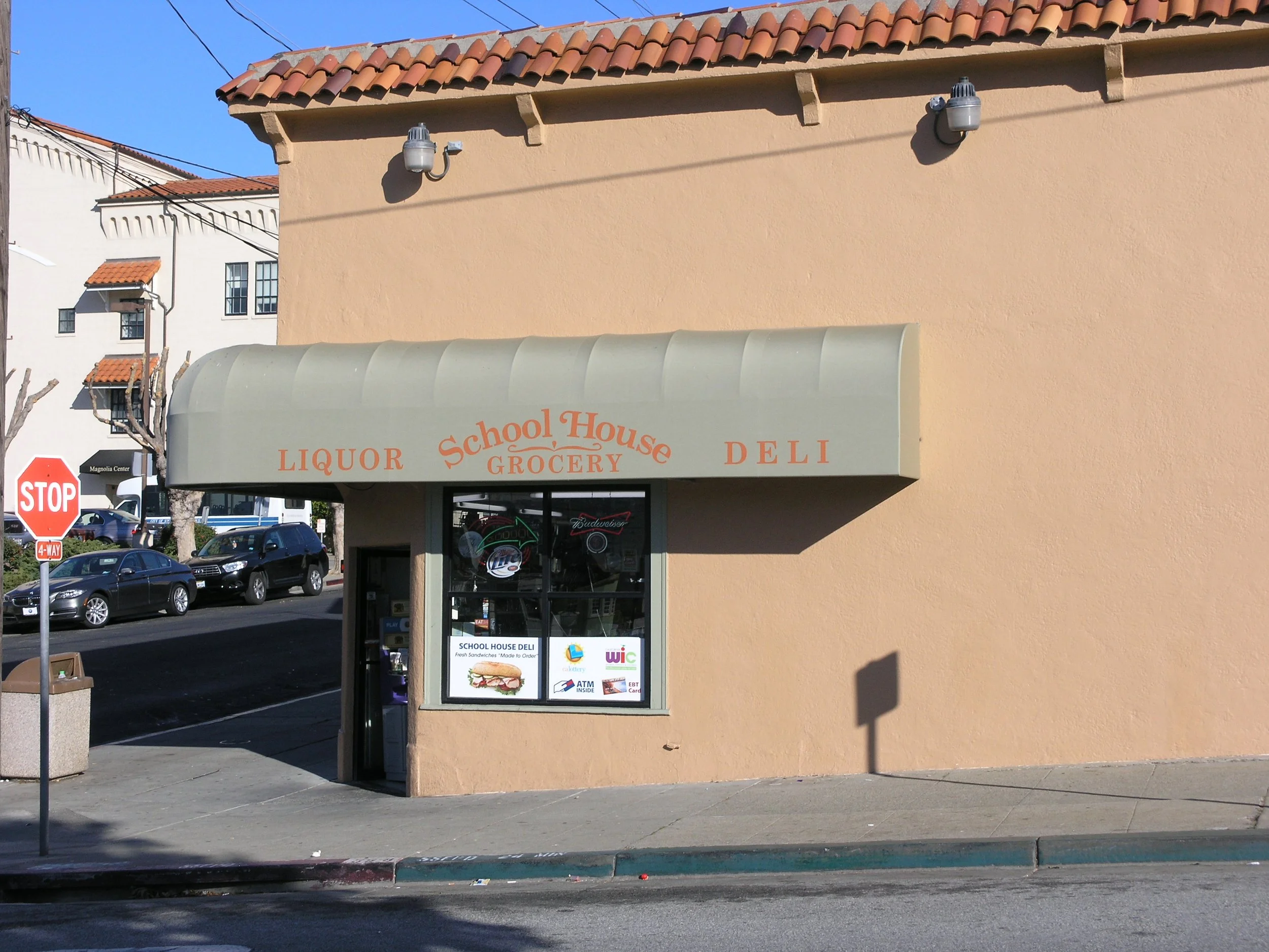 Street view of a beige building with a store named "School House Deli" with a cream-colored awning. Multiple parked cars and a stop sign are visible in the street outside.