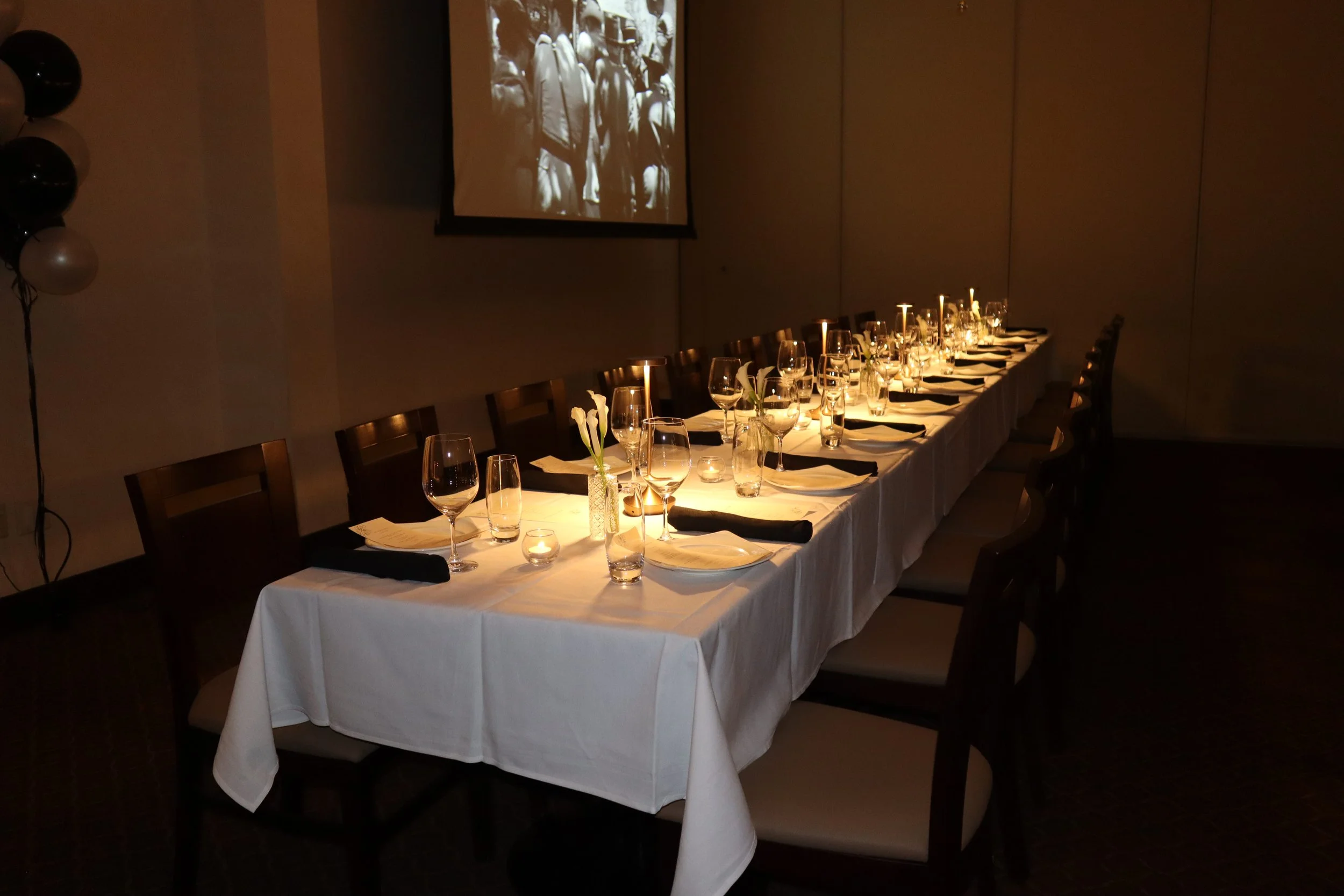 A long banquet table set for a formal dinner with white tablecloths, wine glasses, individual place settings, small flower arrangements, and candles. There are chairs along both sides of the table. In the background, a projection screen displays a bl