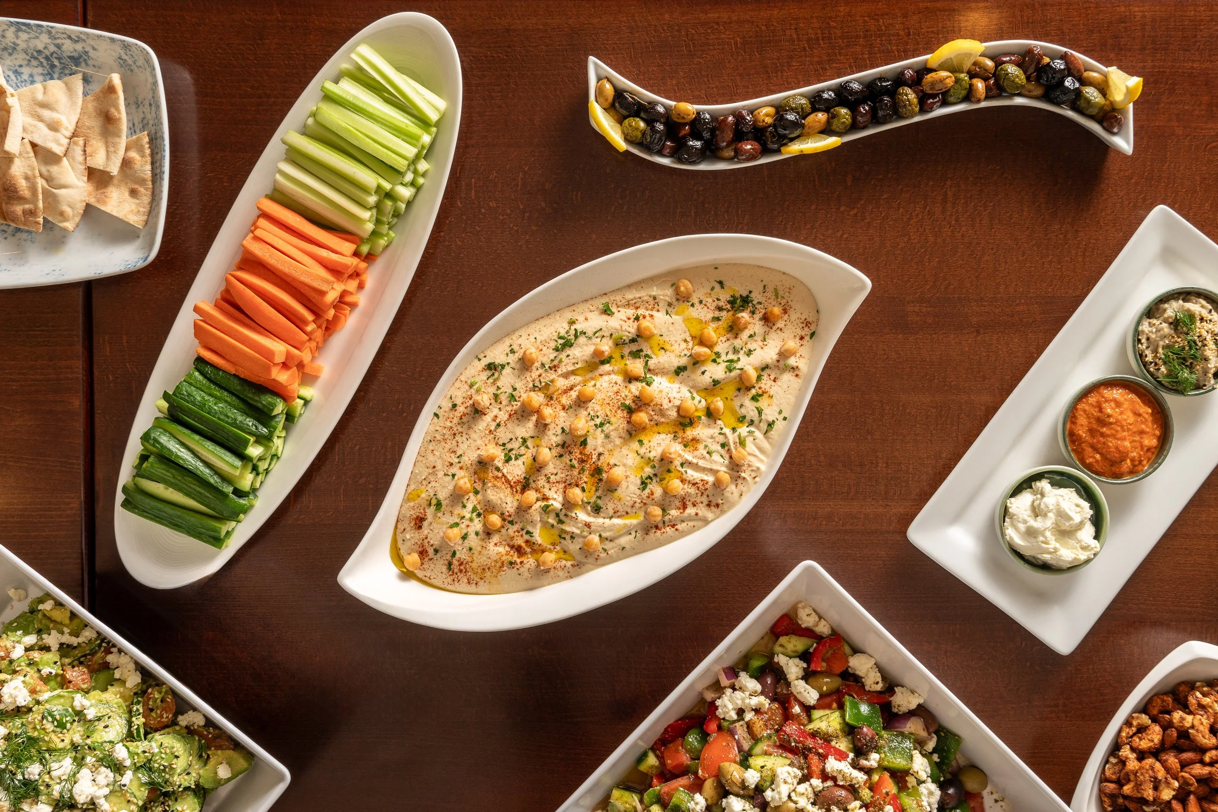 A variety of Mediterranean appetizers on white dishes, including hummus topped with chickpeas and herbs, sliced vegetables, olives, and small bowls of dips and sauces.