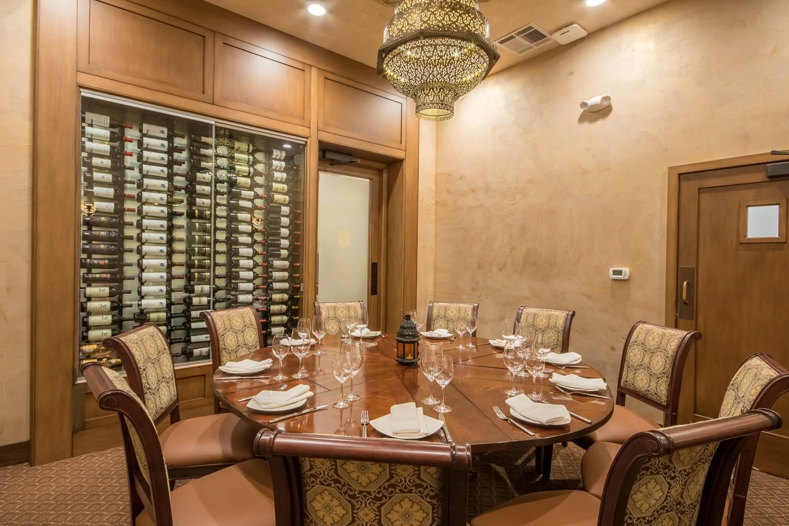 A restaurant dining room with a round wooden table set with wine glasses, plates, and napkins, surrounded by ornate upholstered chairs. There is a large wine rack with bottles behind a glass door, wooden paneled walls, and a decorative chandelier hanging from the ceiling.
