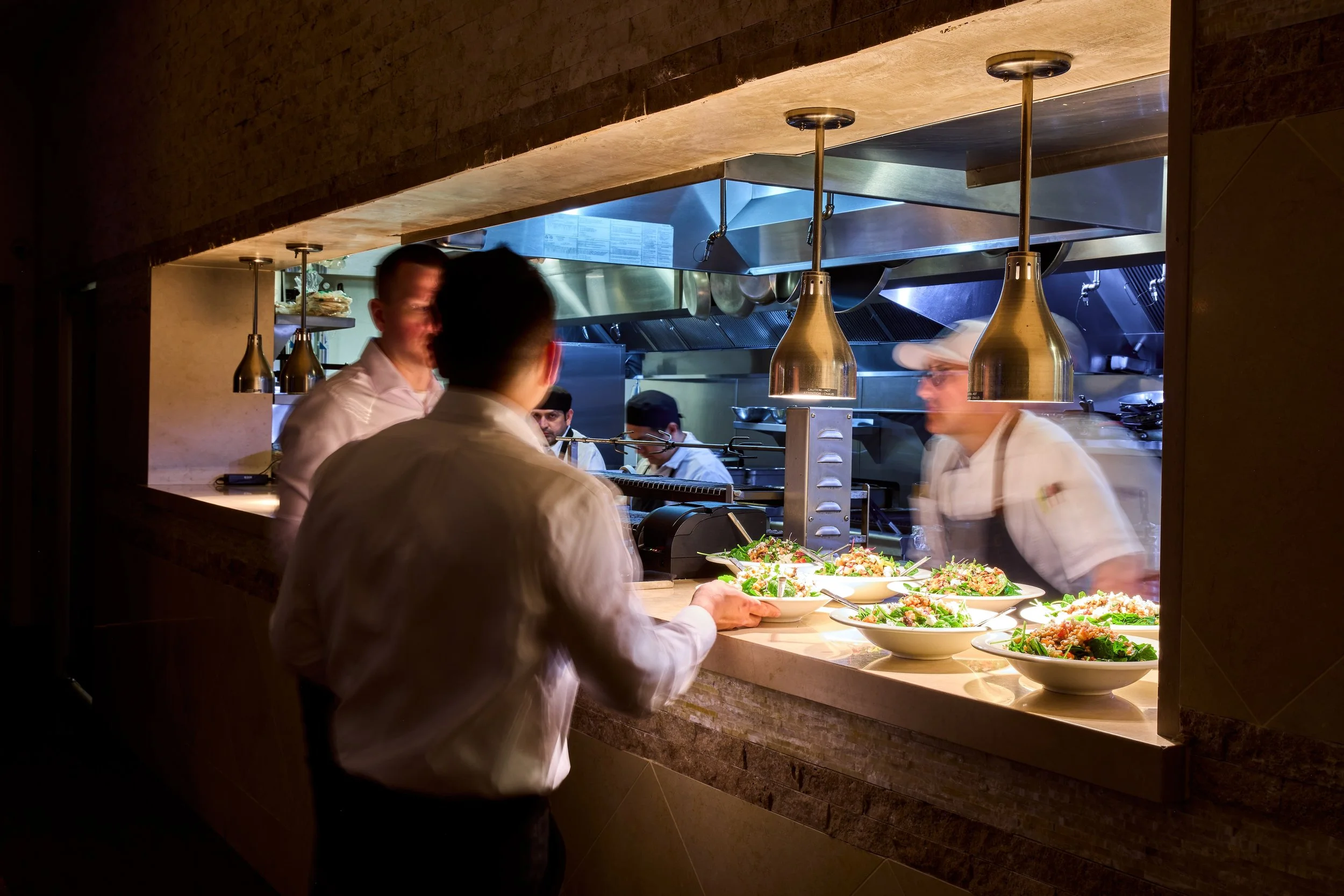 View of a busy restaurant kitchen with chefs preparing food, visible through a window. Plates of salad are on the counter in the foreground.