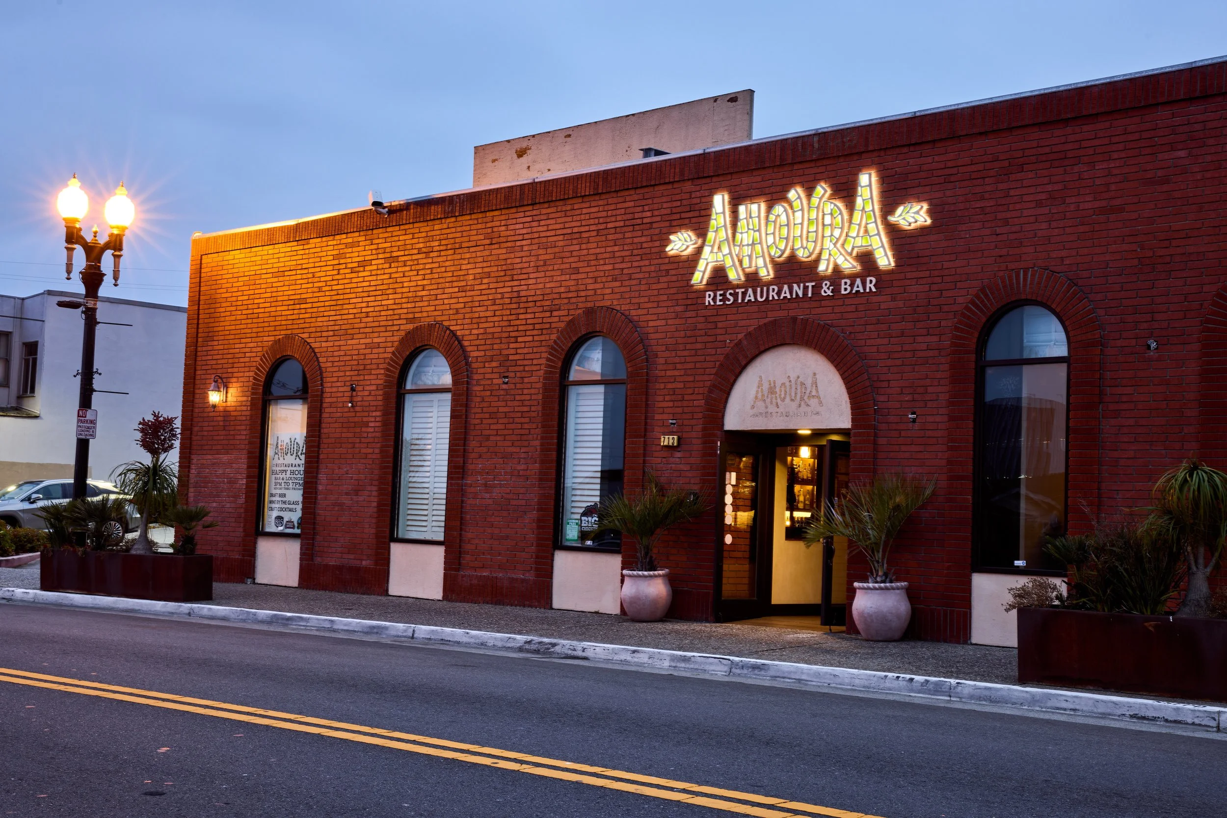 Exterior of a brick restaurant and bar called Amoura, with illuminated sign, arched windows, decorative plants outside, and a streetlamp at dusk.