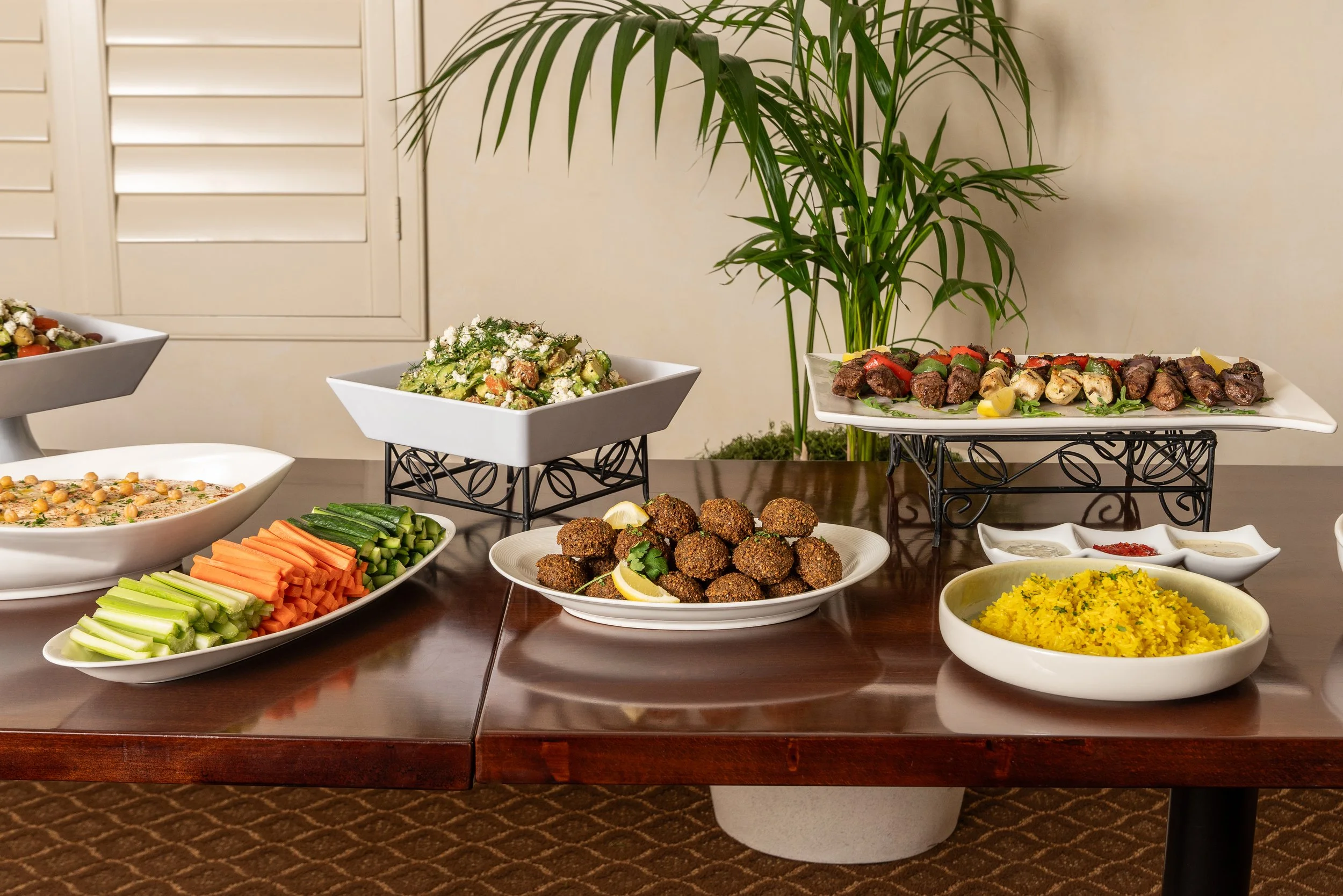 A buffet table with various dishes, including vegetable and meat appetizers, surrounded by green plants in a neutral-colored room.