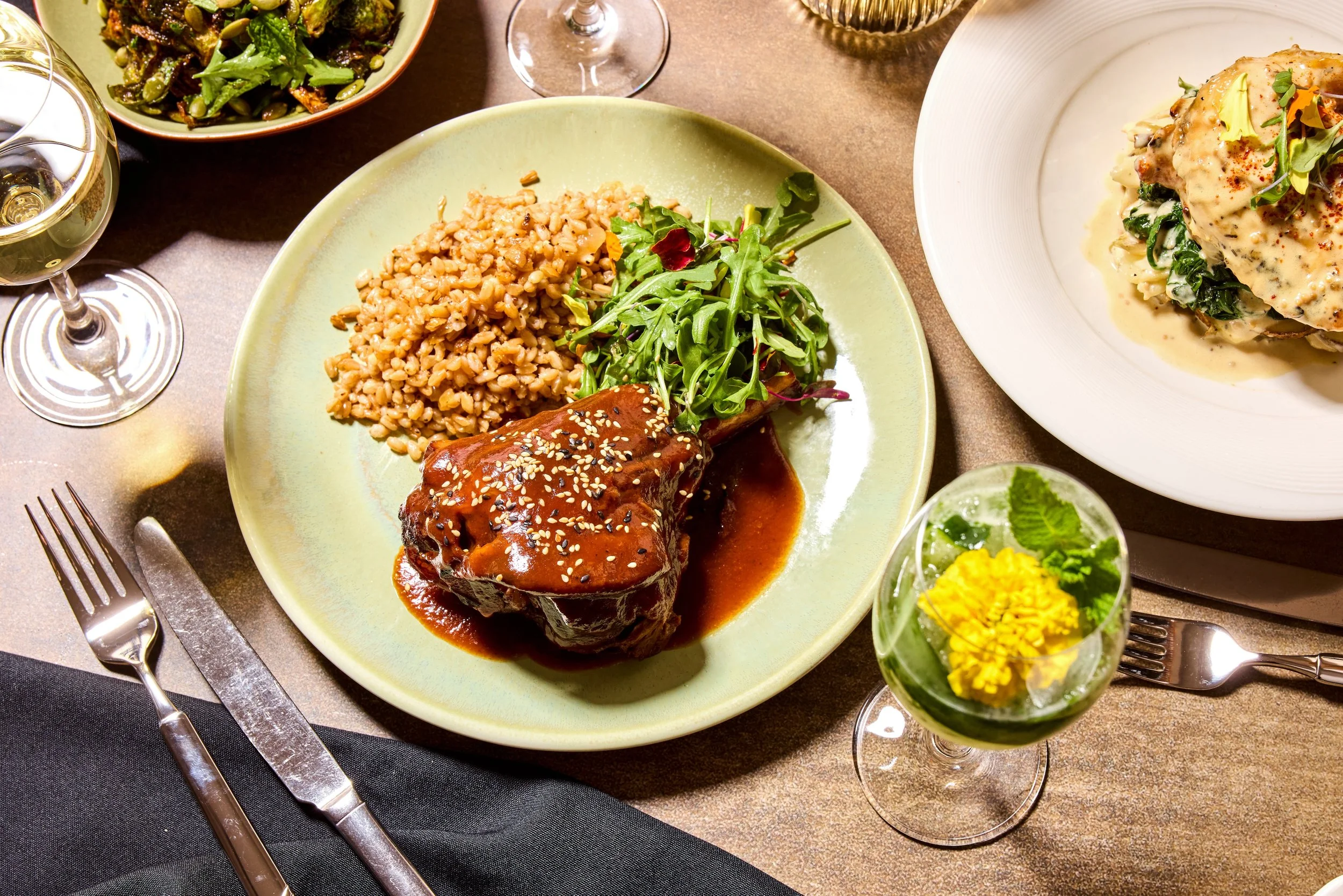 A plate with rice, a glazed meat dish topped with sesame seeds, and a side salad with greens. Surrounding the plate are wine glasses, a spoon, a fork, and a small glass with yellow flowers, garnished with herbs, along with other bowls and glasses on 