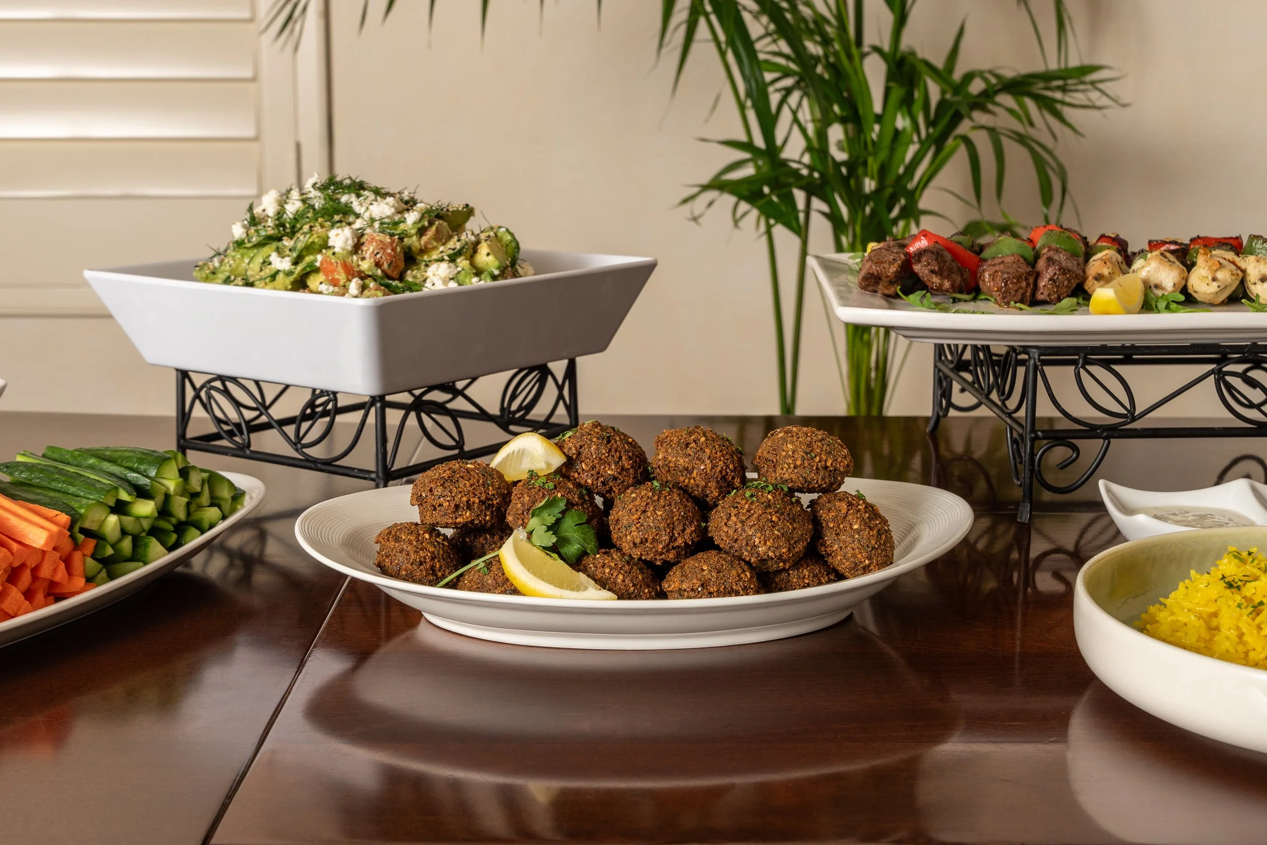 A variety of dishes on a dining table, including a plate of falafel, a tray of meatballs, vegetable skewers with peppers and chicken, a vegetable salad, and fresh sliced vegetables like cucumbers, carrots, and celery.