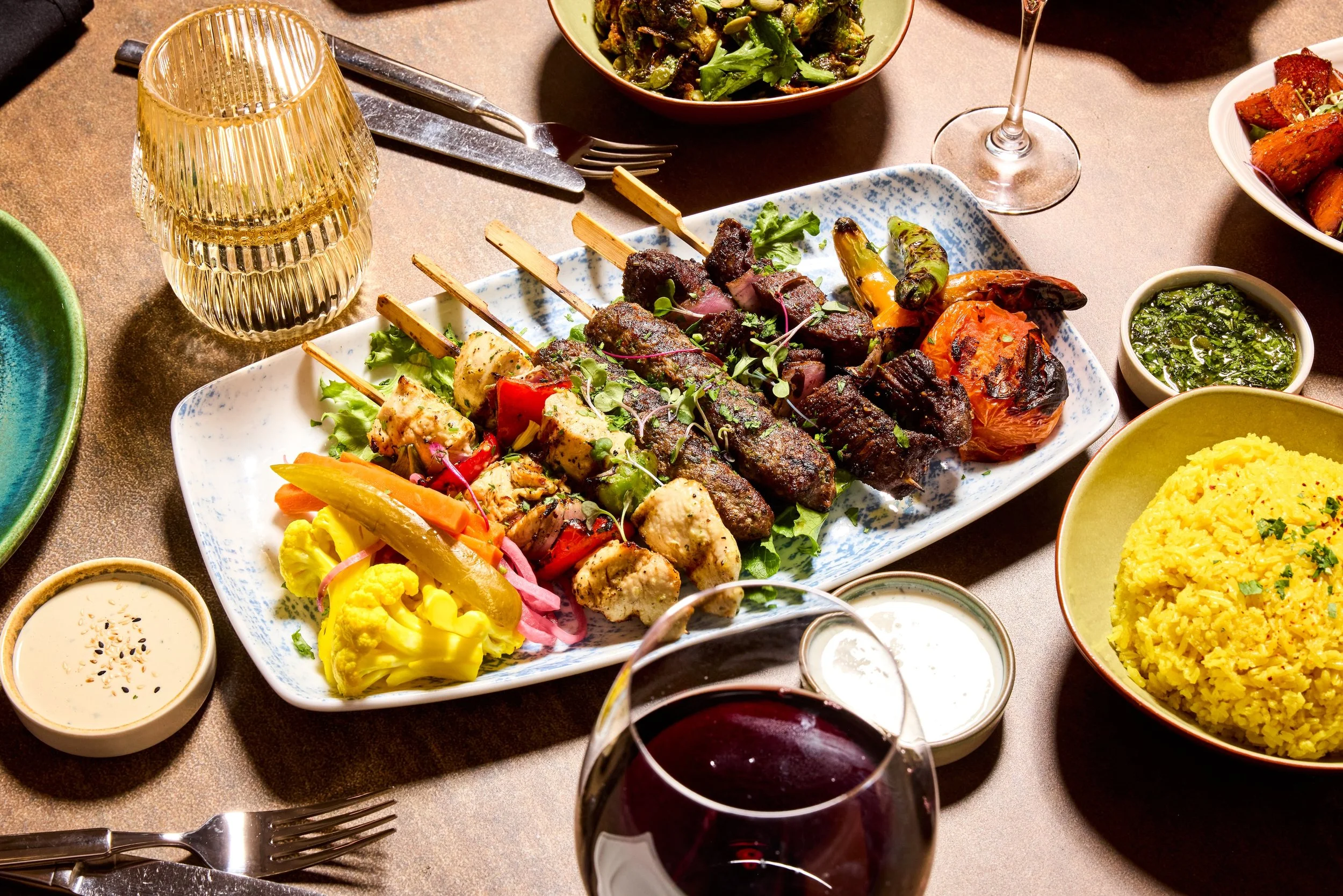 A table with various dishes including skewered meats, vegetables, rice, and multiple bowls of sauces, along with glasses of red wine.