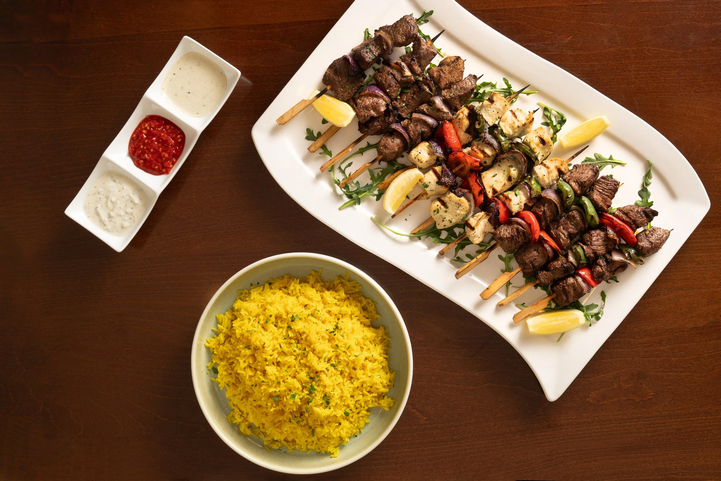 A white rectangular plate with grilled skewers of beef, chicken, and vegetables, garnished with lemon wedges. Next to the plate, a long white dish with three compartments holding sauces. A bowl of yellow rice and a bowl of yellow saffron rice are als