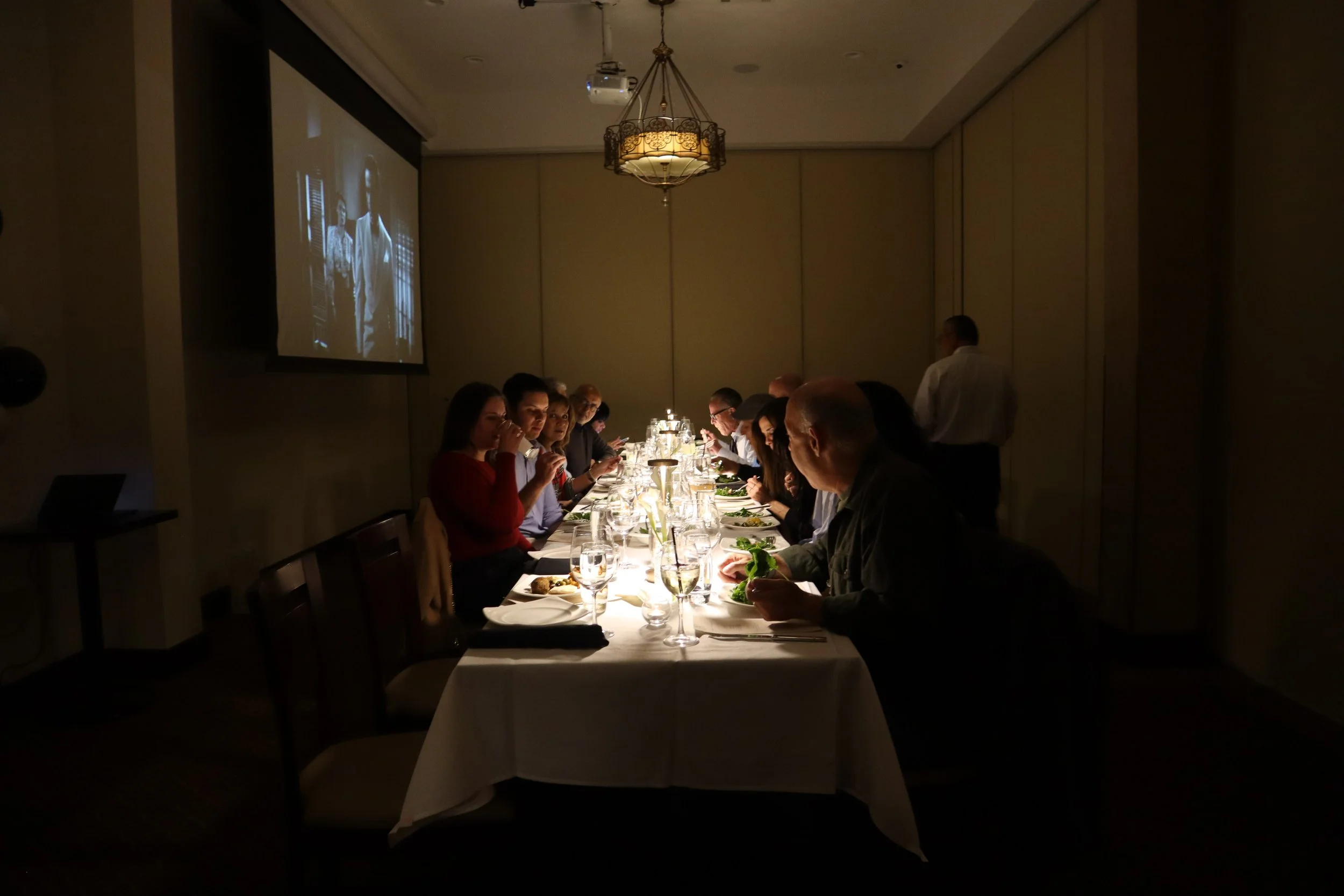 A group of people seated at a long dining table in a dimly lit restaurant or banquet hall, with a chandelier hanging from the ceiling. There is a large screen on the wall displaying a black-and-white image, and a waiter is standing near the wall.