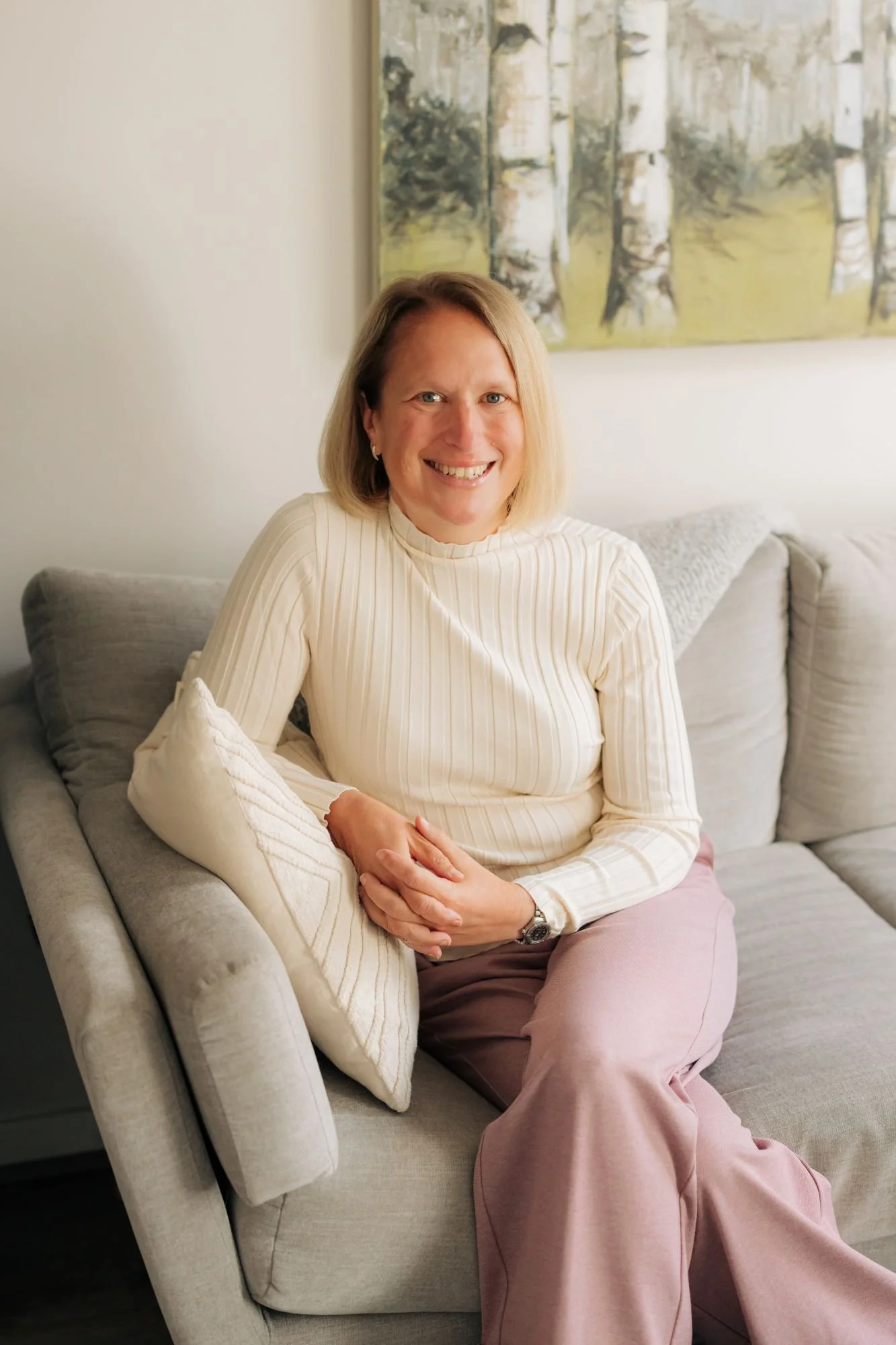 A smiling woman with shoulder-length blond hair, wearing a cream ribbed turtleneck sweater and light pink pants, sitting on a light gray sofa with cream and beige pillows, in a room with white walls and a modern abstract painting of birch trees in the background.