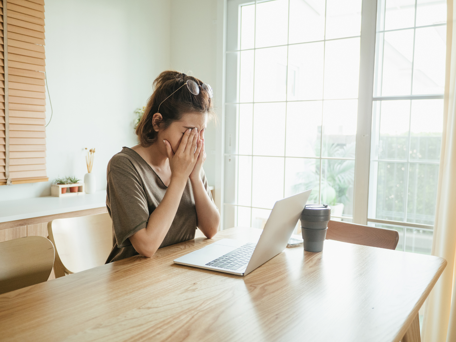 Woman sitting at a wooden table with a closed laptop and a travel mug, covering her face with her hands in a bright room with large windows.